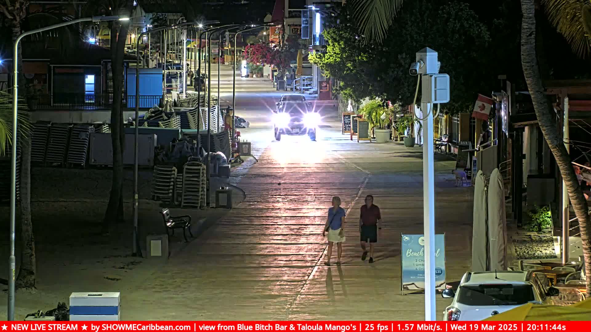A nighttime street scene shows a paved walkway lined with beach chairs and buildings, with a vehicle's headlights illuminating the path as two pedestrians walk towards the camera.
