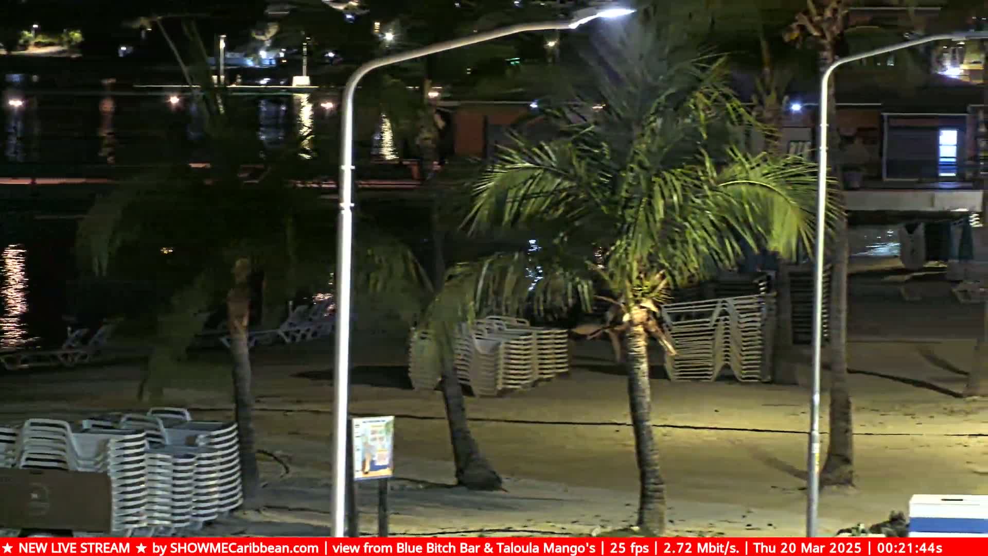 A nighttime beach scene shows calm water, palm trees, and stacks of beach chairs under artificial light.