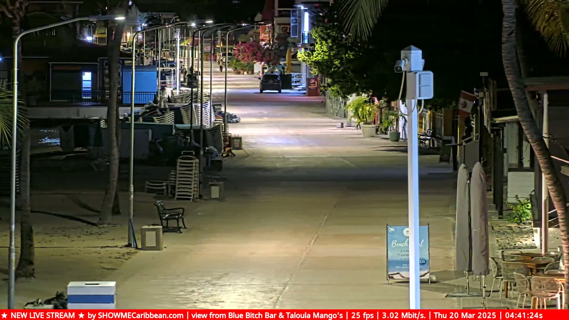 A nighttime view of a mostly empty boardwalk next to a beach, with palm trees, streetlights, and some outdoor furniture.