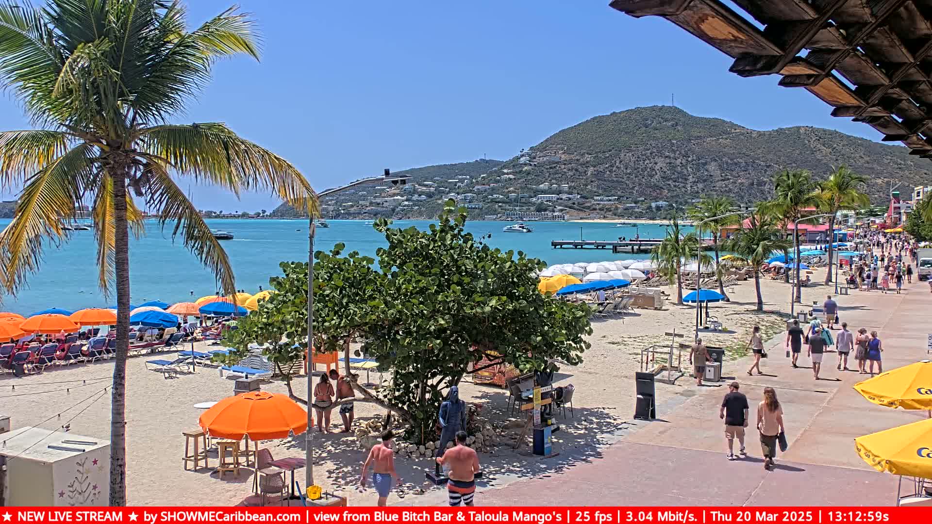 A sunny beach scene shows a sandy shore lined with colorful umbrellas and beachgoers, a boardwalk with pedestrians, and a calm ocean with boats in the distance, backed by a hillside town under a clear sky.