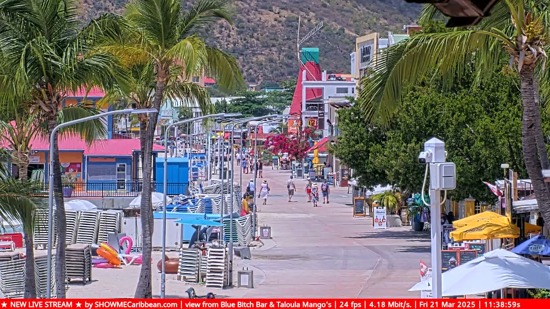 A sunny boardwalk scene shows pedestrians strolling past colorful buildings and beach chairs stacked near the water's edge, under a clear sky.