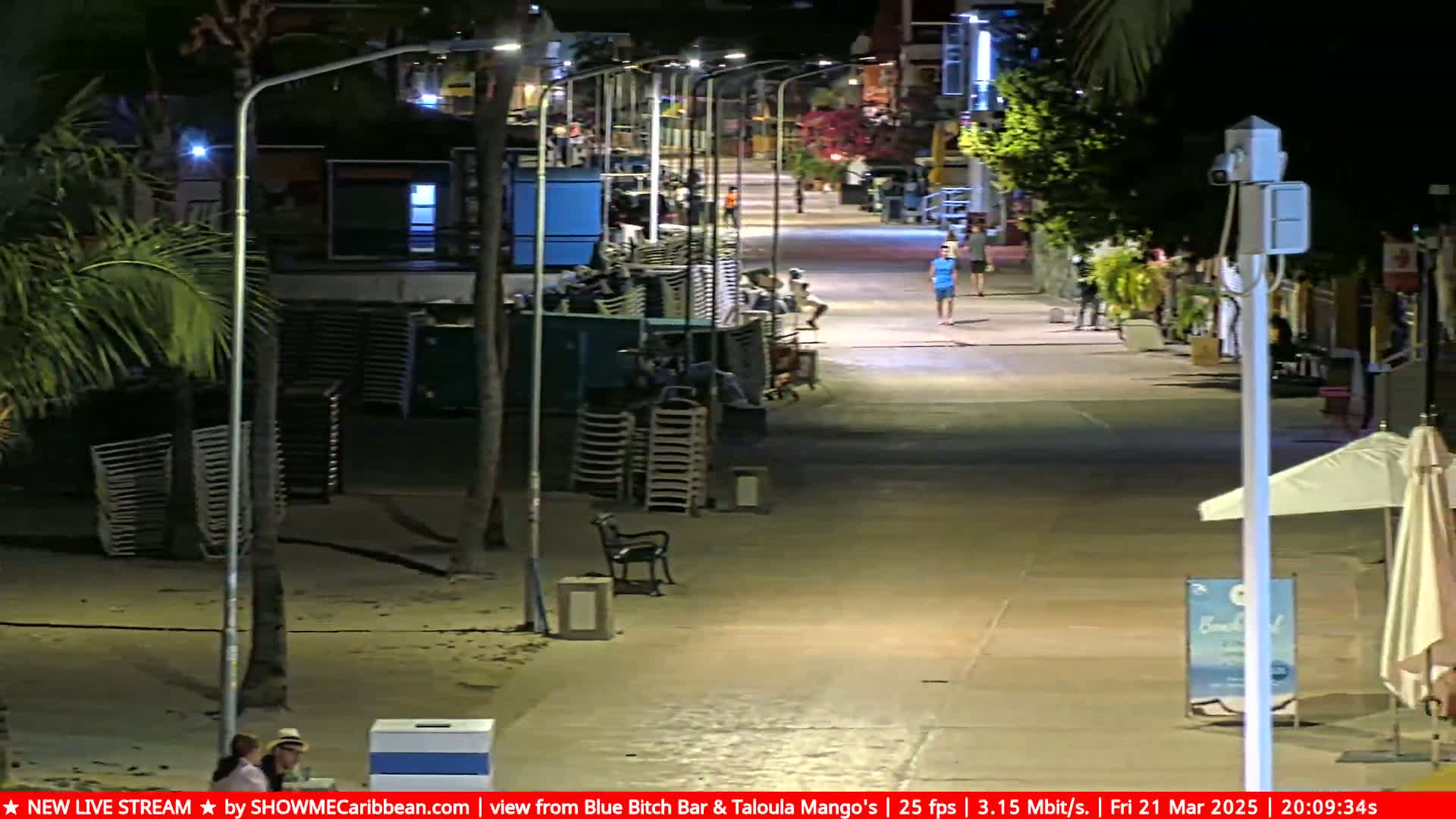 A nighttime view of a mostly empty beachside boardwalk with stacked beach chairs and a few people walking, under clear skies.