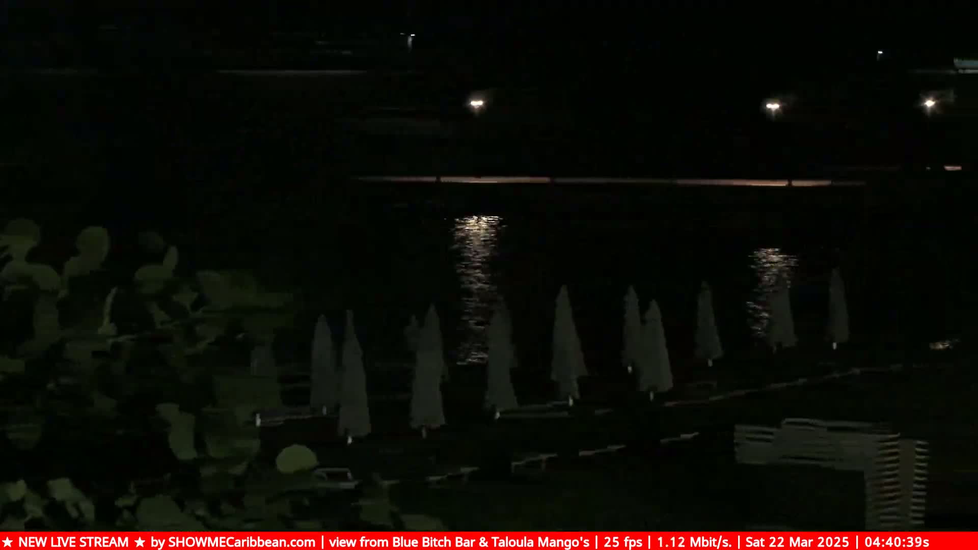 A nighttime view of a calm body of water reflecting lights, with closed beach umbrellas and lounge chairs on the shore.