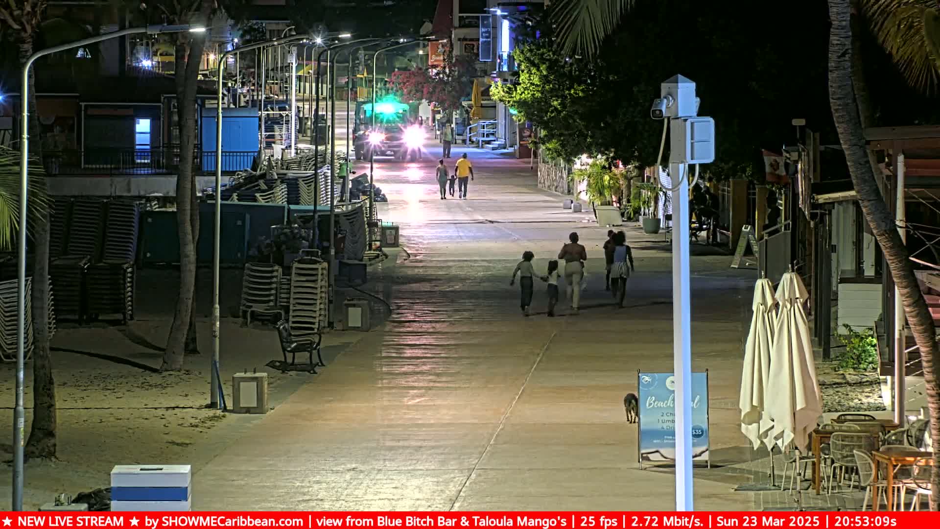 At night, several people walk along a paved beachfront path lined with palm trees and buildings, under clear skies.