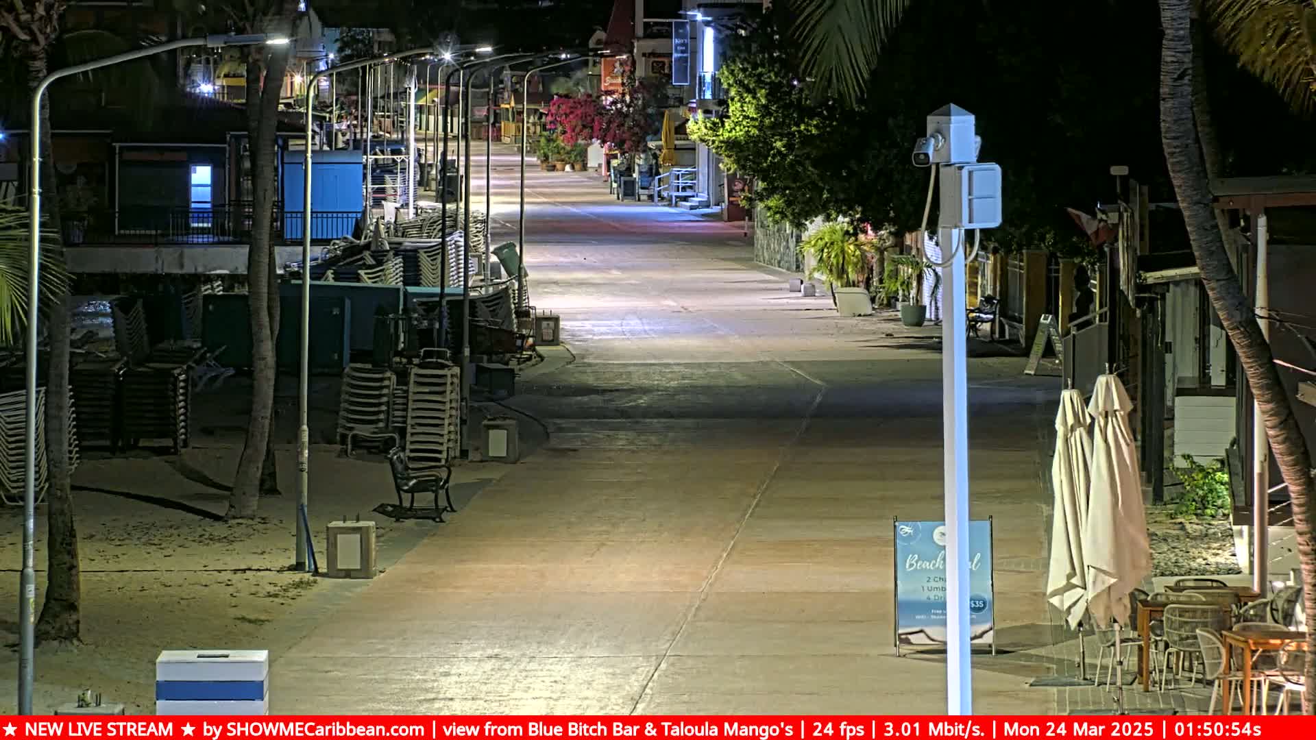 A mostly empty, brightly lit boardwalk at night, lined with stacked beach chairs and some outdoor seating areas under palm trees.