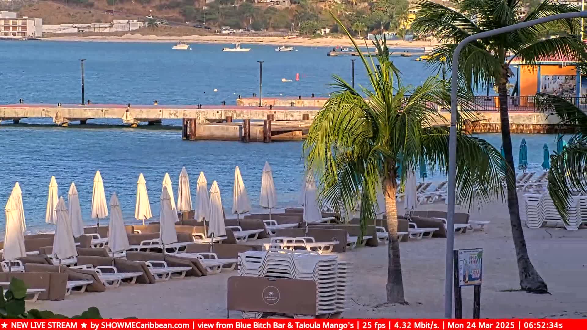 A sandy beach with numerous lounge chairs and umbrellas under a partly sunny sky overlooks a calm body of water with a pier in the distance.