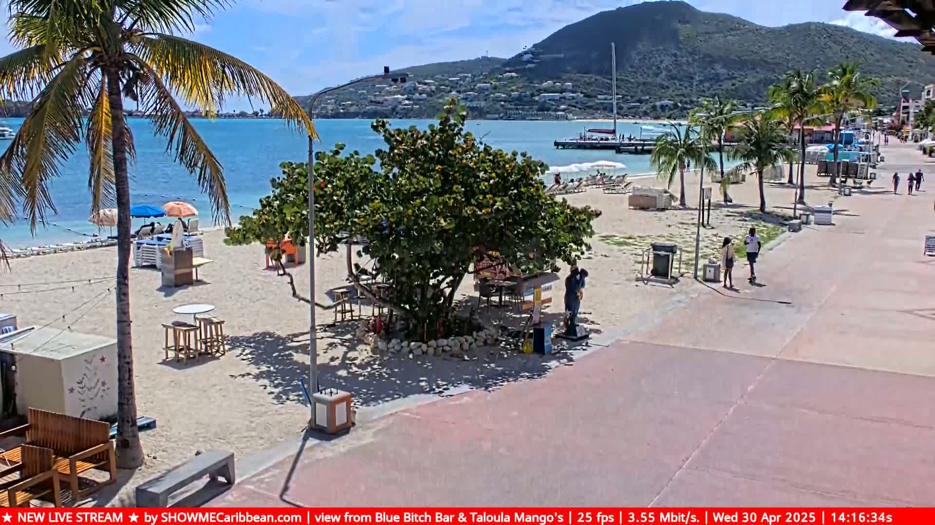 A sunny beach scene shows a sandy shore with beach umbrellas, lounge chairs, and some people walking on a paved boardwalk, all under a bright blue sky with a calm sea and distant hills.
