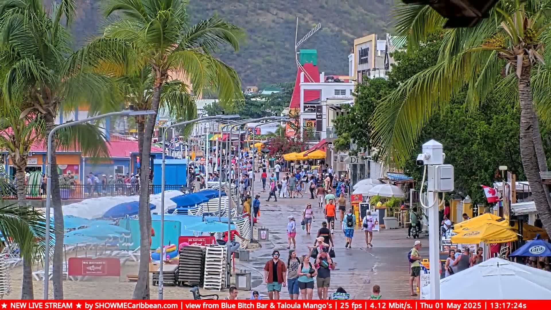 A wet boardwalk lined with shops and restaurants is crowded with people on a cloudy day, with beach umbrellas and lounge chairs visible on the sand.