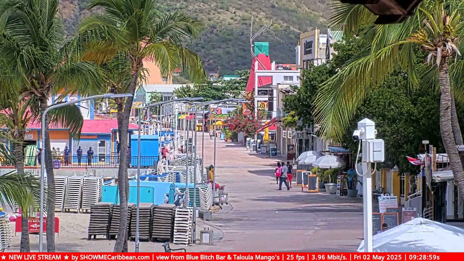 A sunny boardwalk lined with colorful shops and palm trees, with stacks of beach chairs on the sand to the side.