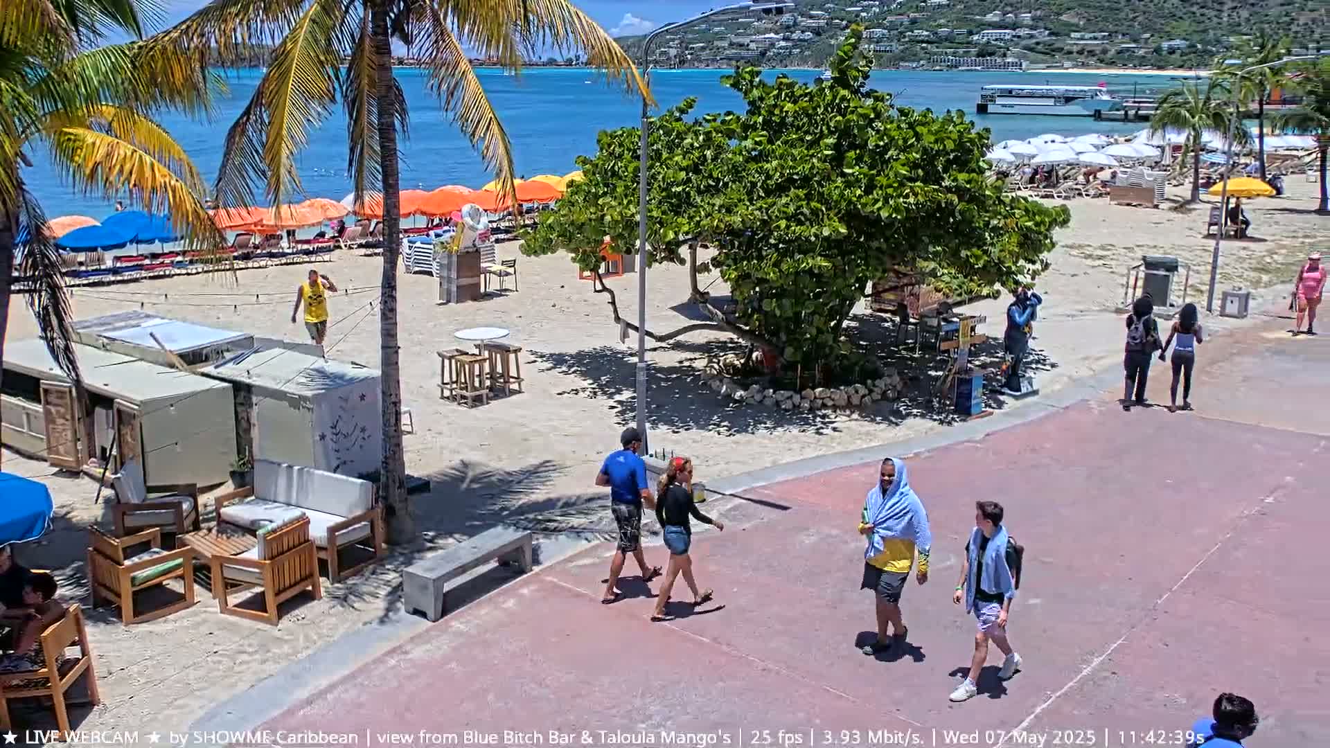 A sunny beach scene shows people walking on a paved area near a sandy beach with colorful umbrellas, beach chairs, and some outdoor furniture.