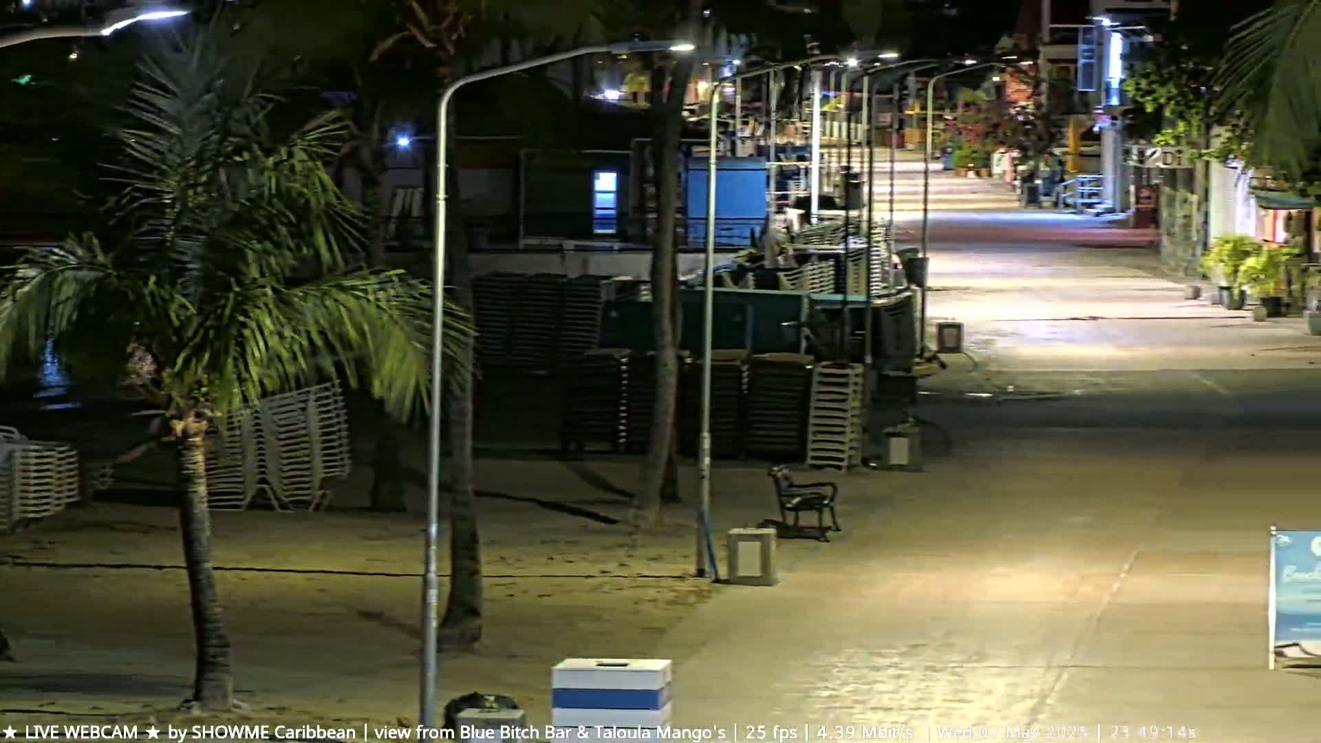 A nighttime view of a mostly deserted beachside street lined with palm trees, lampposts, and stacks of beach chairs.