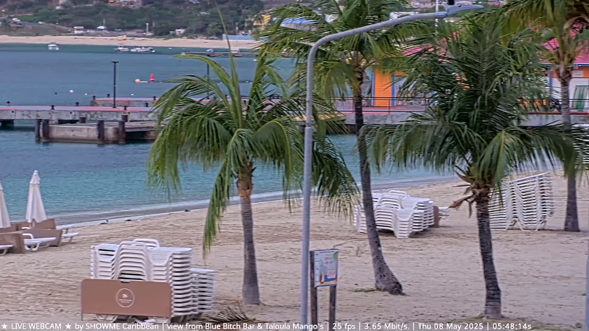A sandy beach with palm trees and stacks of white lounge chairs is visible next to calm, turquoise water and a pier, under an overcast sky.