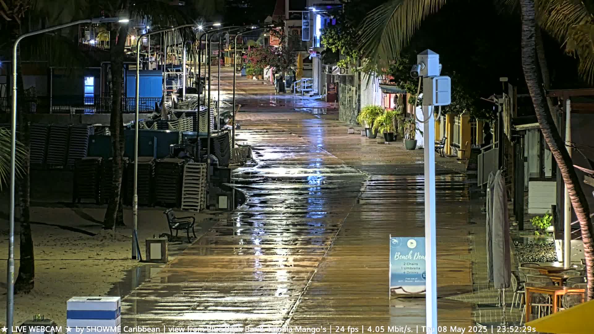 A wet boardwalk at night, lined with closed shops and stacked beach chairs, is illuminated by streetlights.