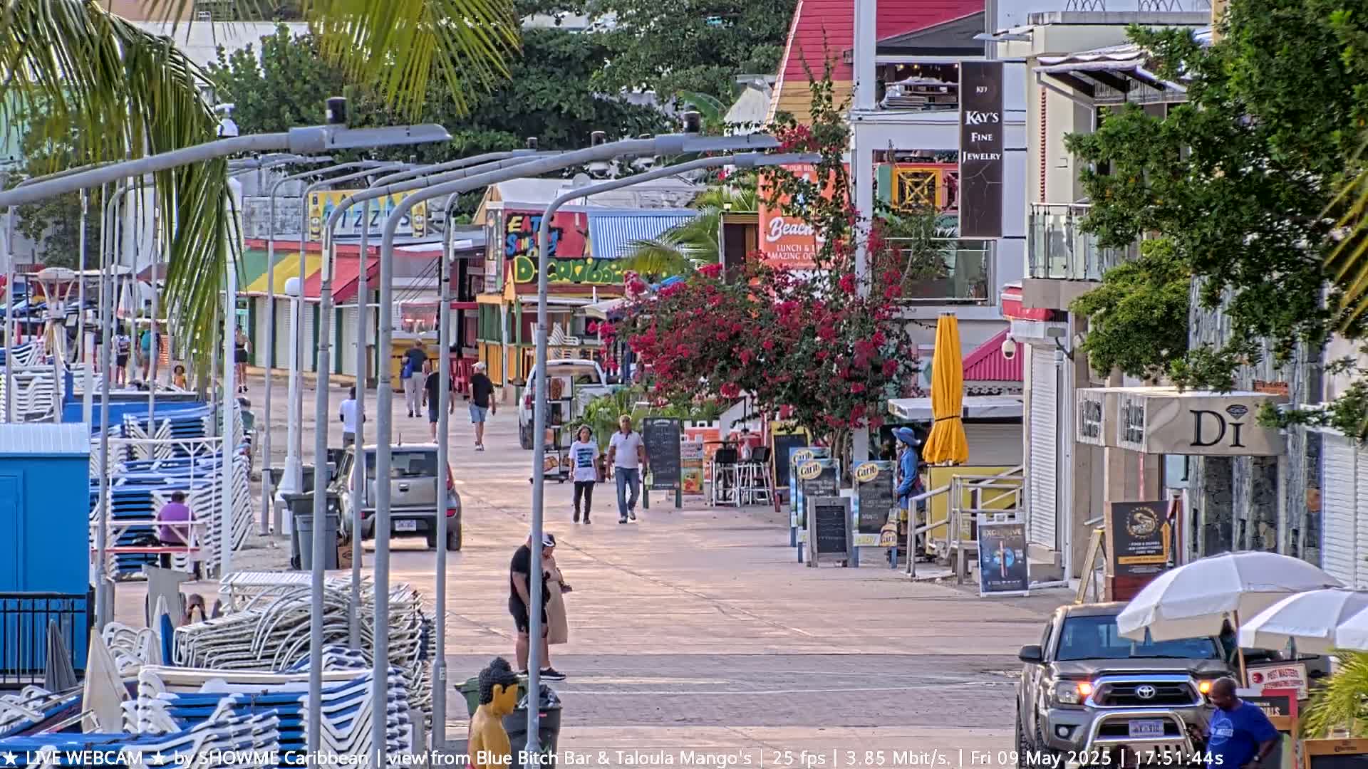 A sunny street scene shows a pedestrian walkway lined with colorful shops and restaurants, beach chairs stacked nearby, and people walking along the street.