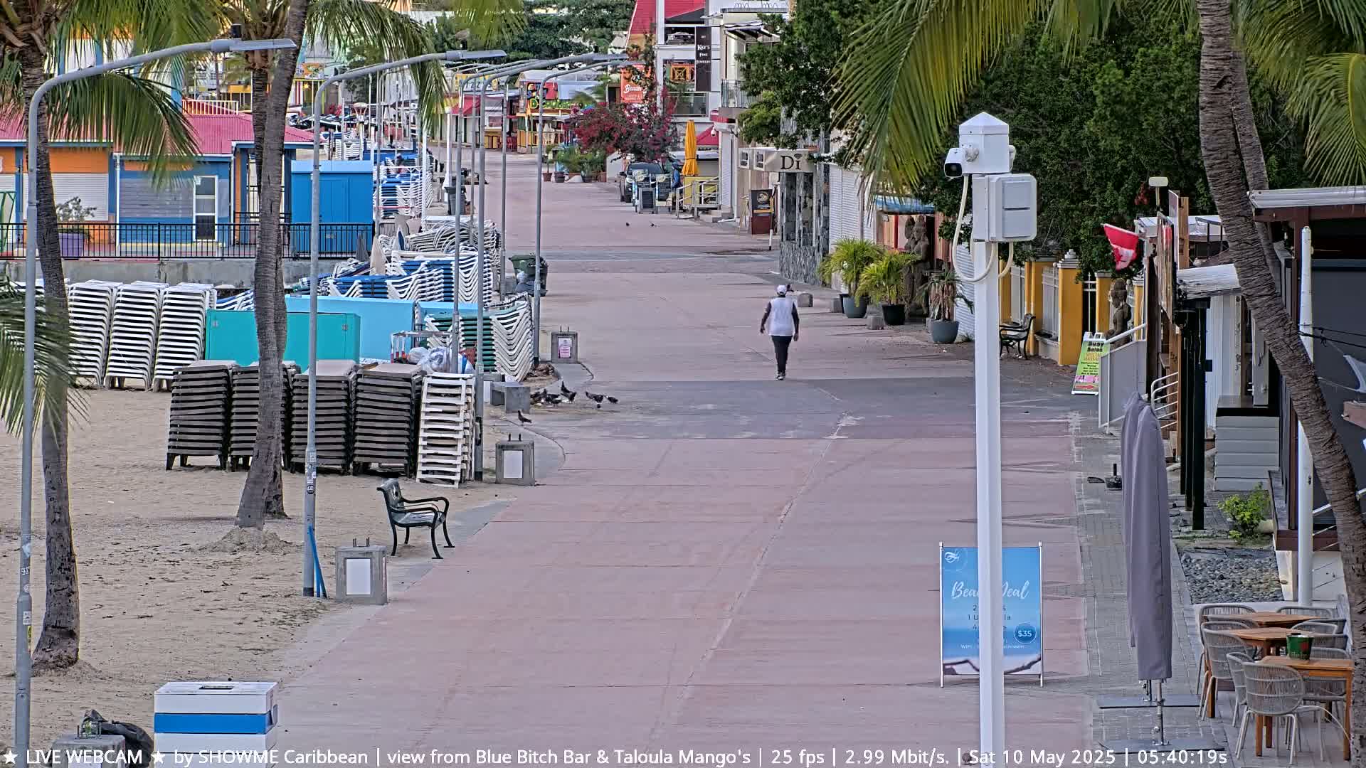 A person walks down a paved walkway lined with stacked beach chairs, palm trees, and shops, under a partly cloudy sky.