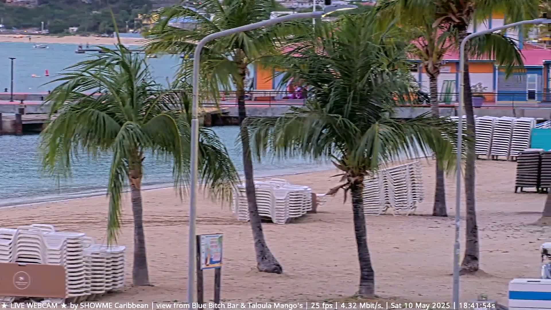 A sandy beach with palm trees and stacks of lounge chairs is visible, next to calm, blue water under an overcast sky.