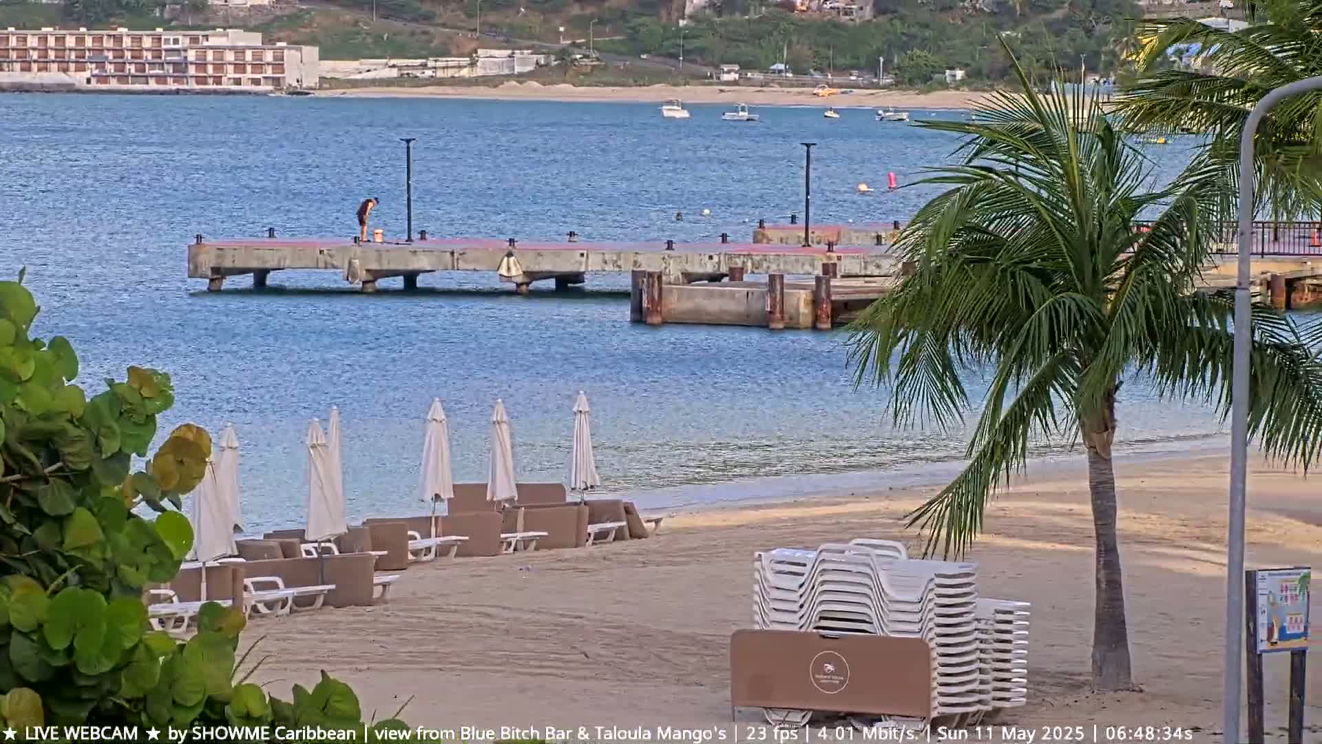 A person stands on a pier extending into calm blue water near a sandy beach with lounge chairs and umbrellas under a partly sunny sky.