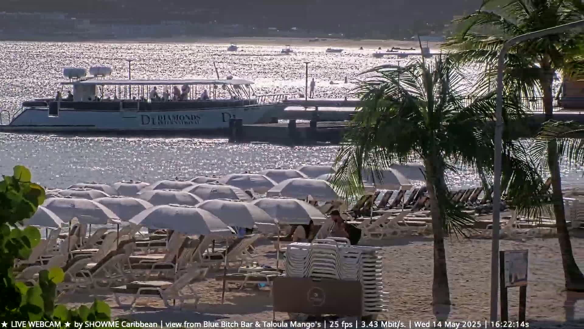 A passenger boat is docked near a sandy beach with numerous white umbrellas and lounge chairs under a sunny sky.