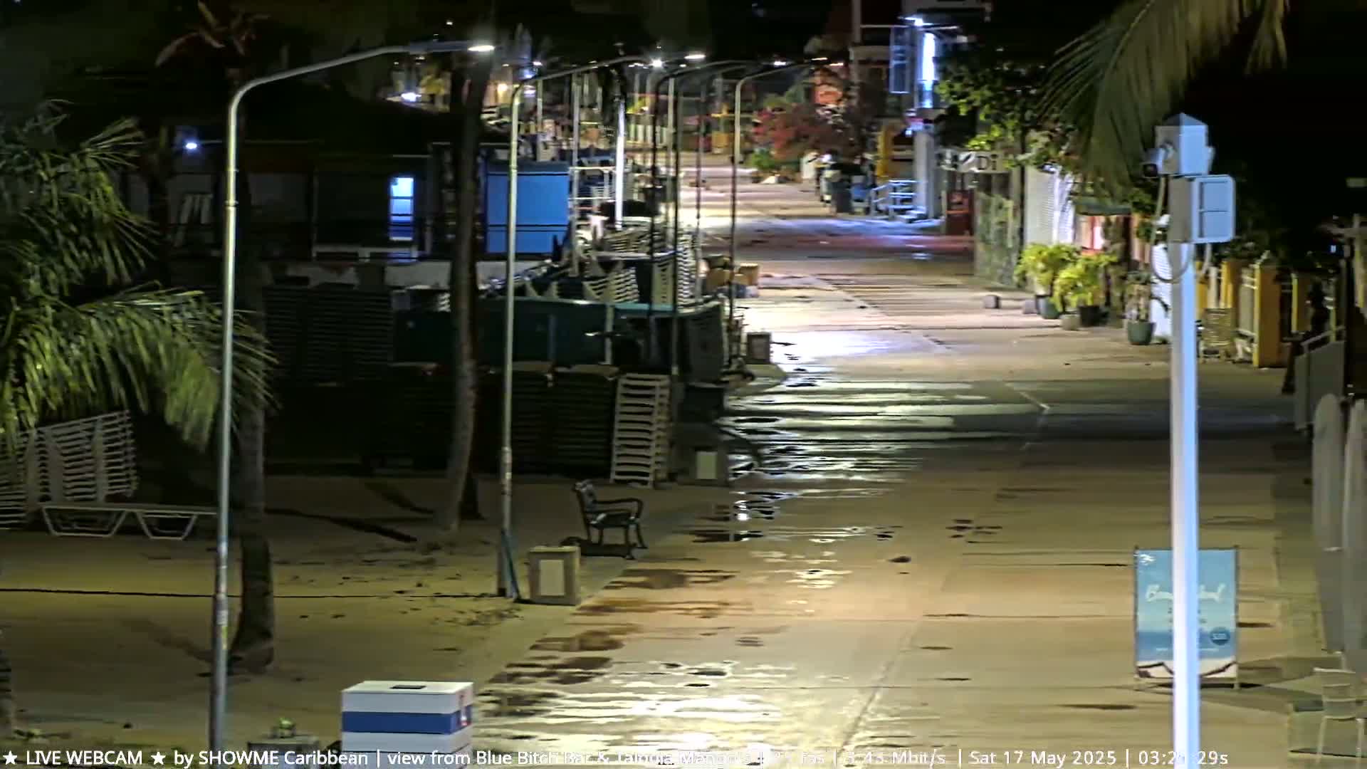 A mostly empty beachside walkway at night, wet from recent rain, is lined with lampposts and stacked beach chairs.