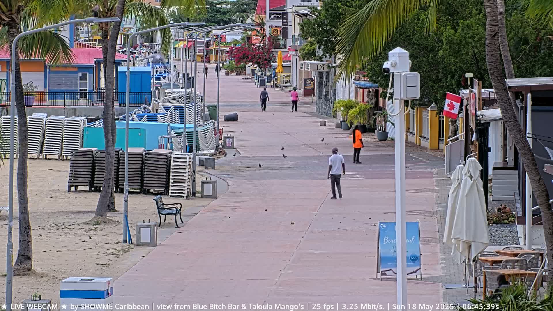 A mostly empty paved walkway lined with shops and stacked beach chairs stretches toward a beach under a partly cloudy sky.