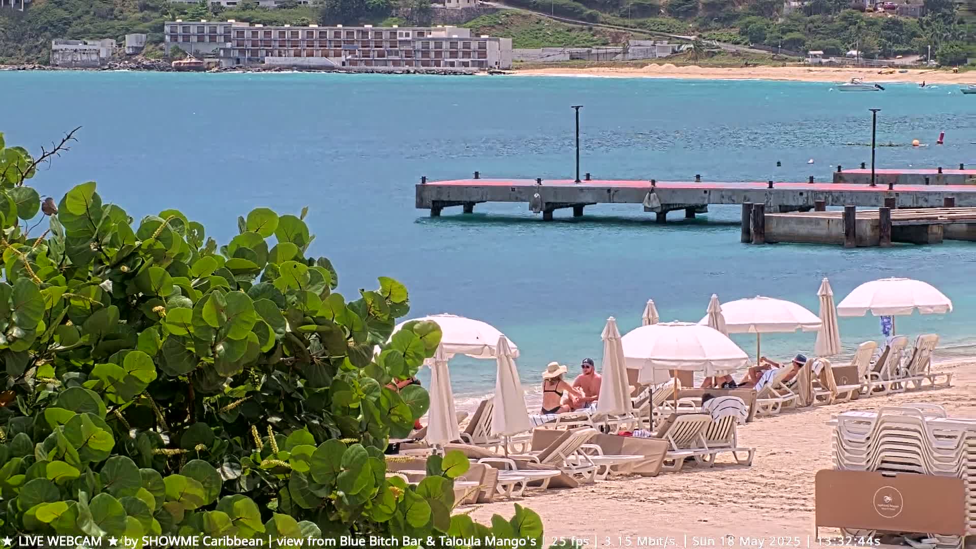 A beach scene with turquoise water, a concrete pier, a multi-story building in the background, and several people relaxing under white umbrellas on the sandy shore.
