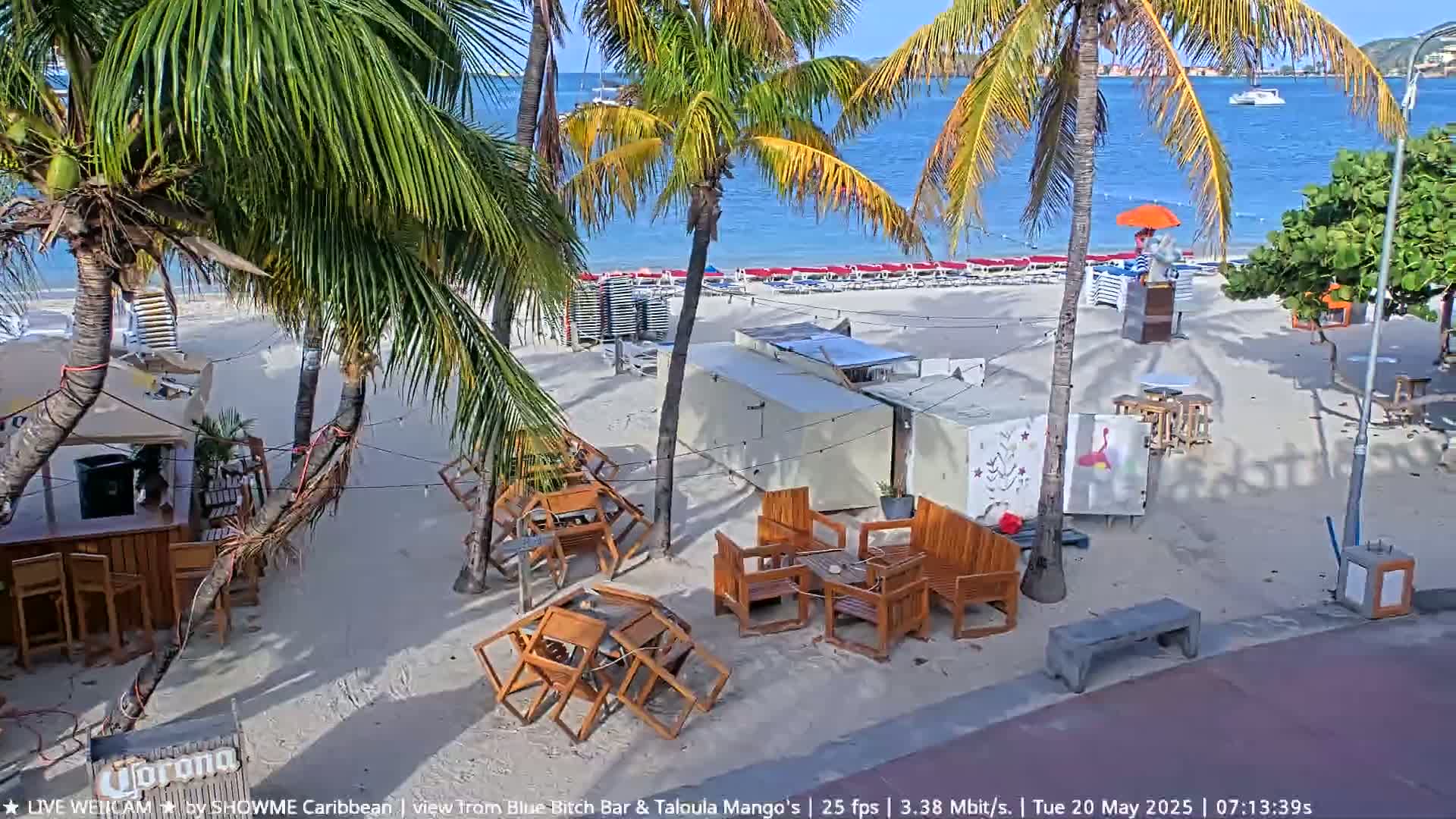 A sandy beach with palm trees, outdoor furniture, and a calm ocean under a sunny sky.