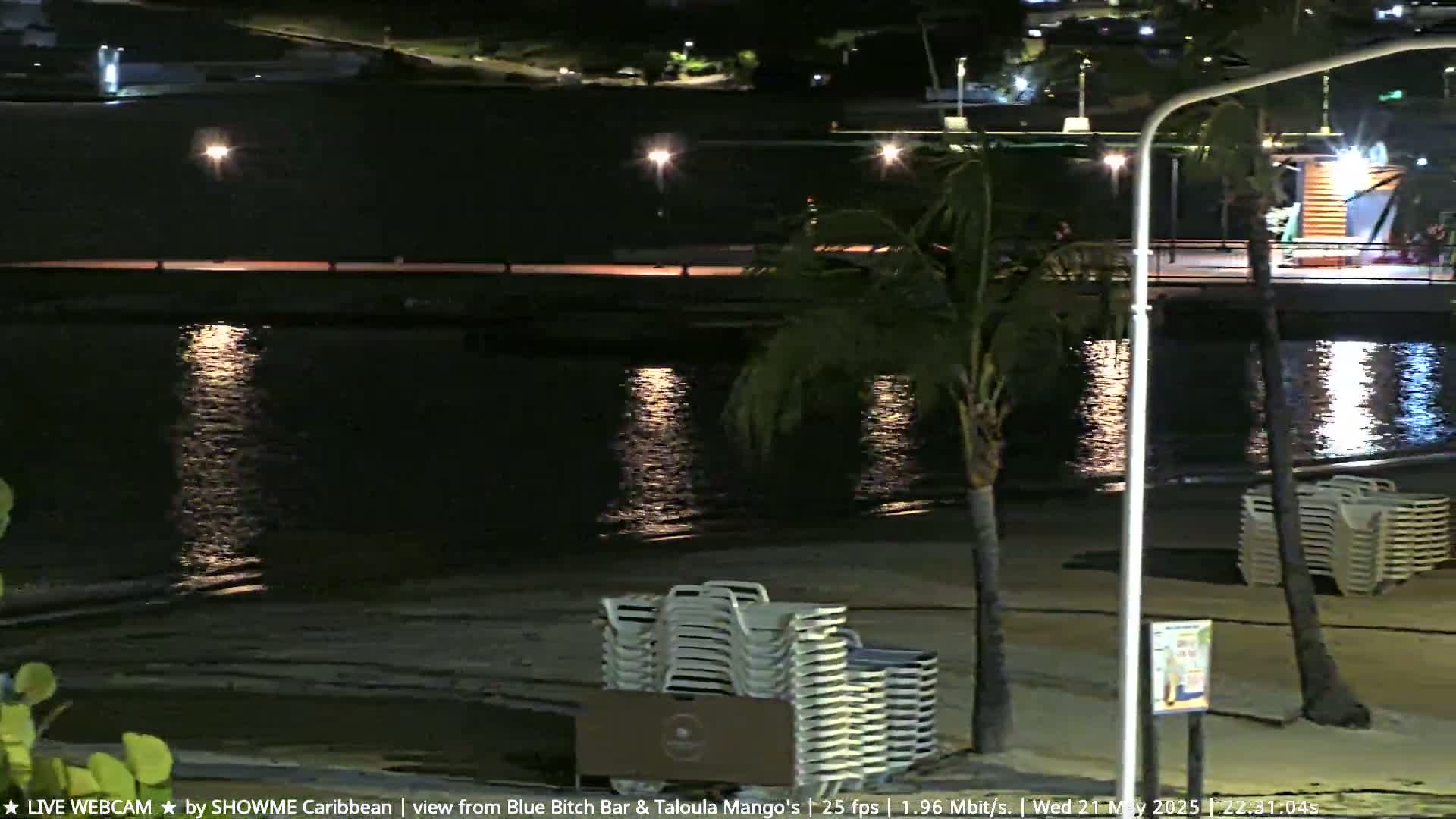A nighttime beach scene shows calm water reflecting lights, stacked beach chairs on the sand, and palm trees under a clear, dark sky.