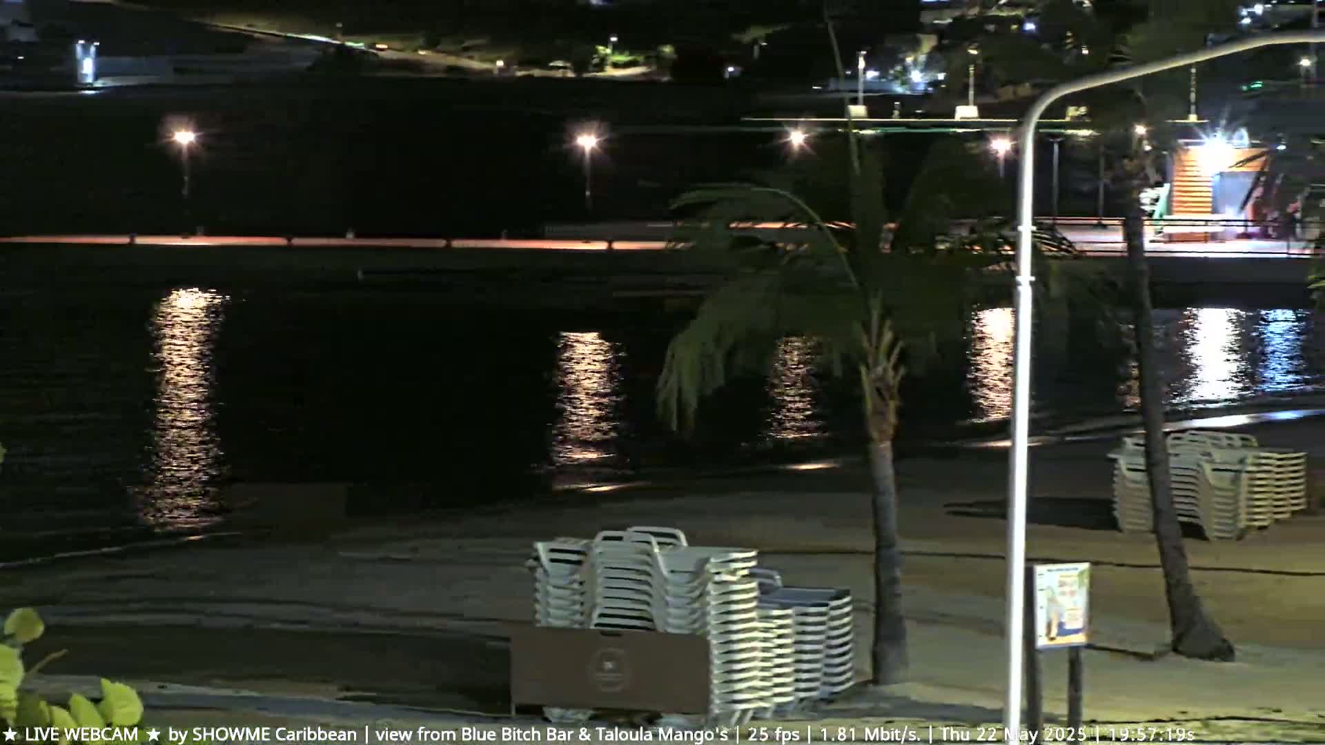 A nighttime beach scene shows calm water reflecting lights from a nearby boardwalk, with stacks of beach chairs on the sand next to palm trees.