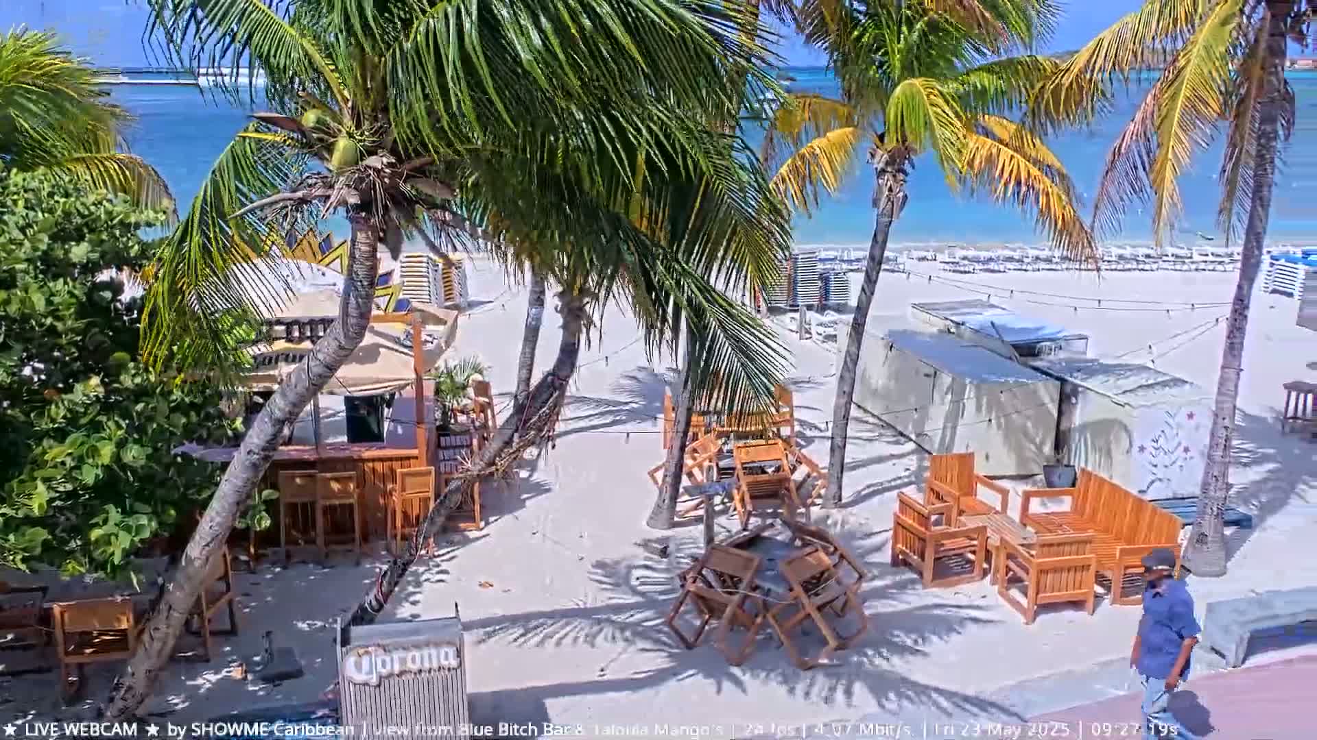 A beach bar with wooden furniture sits on a white sand beach under a sunny sky, with palm trees and turquoise water visible in the background.