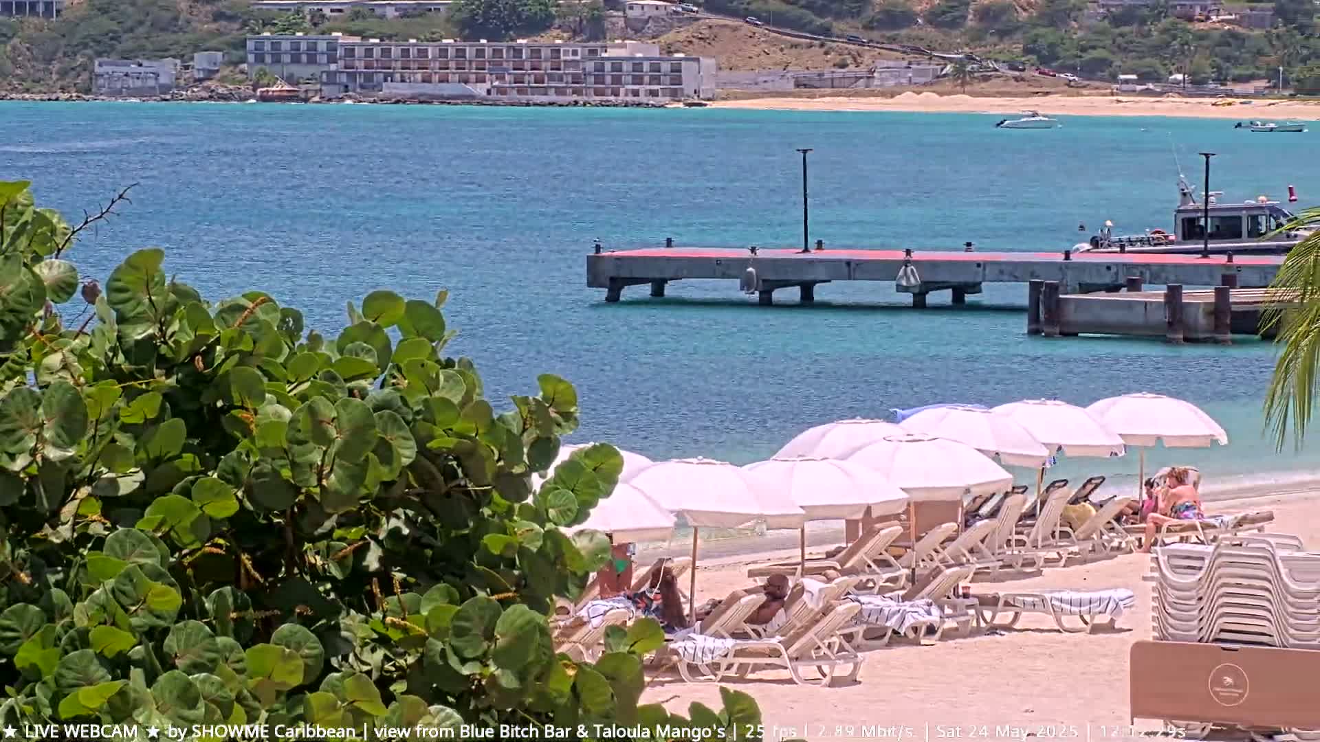 A sandy beach with white umbrellas and lounge chairs overlooks a calm, turquoise ocean where a pier and a small boat are visible, with a building in the distance under a sunny sky.