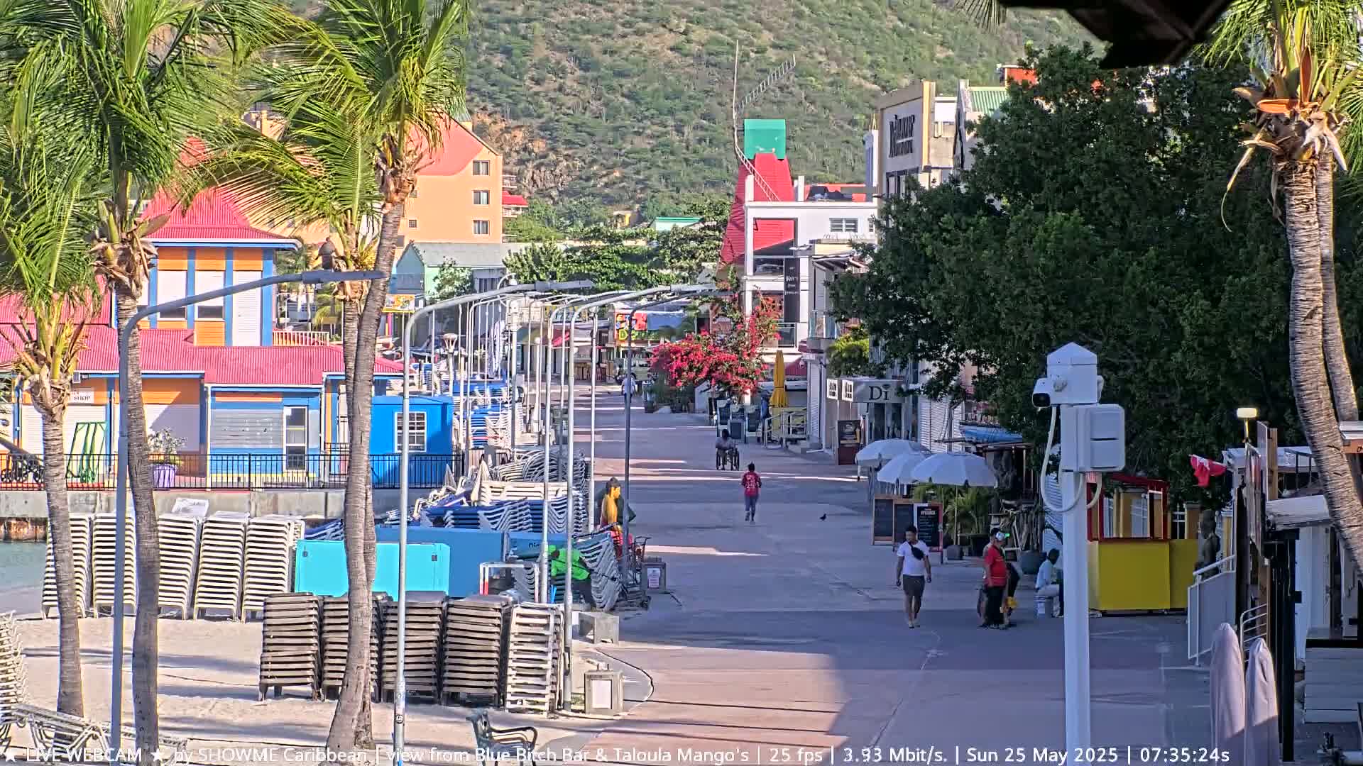 A sunny waterfront boardwalk lined with colorful buildings and stacked beach chairs, with a few pedestrians strolling along.