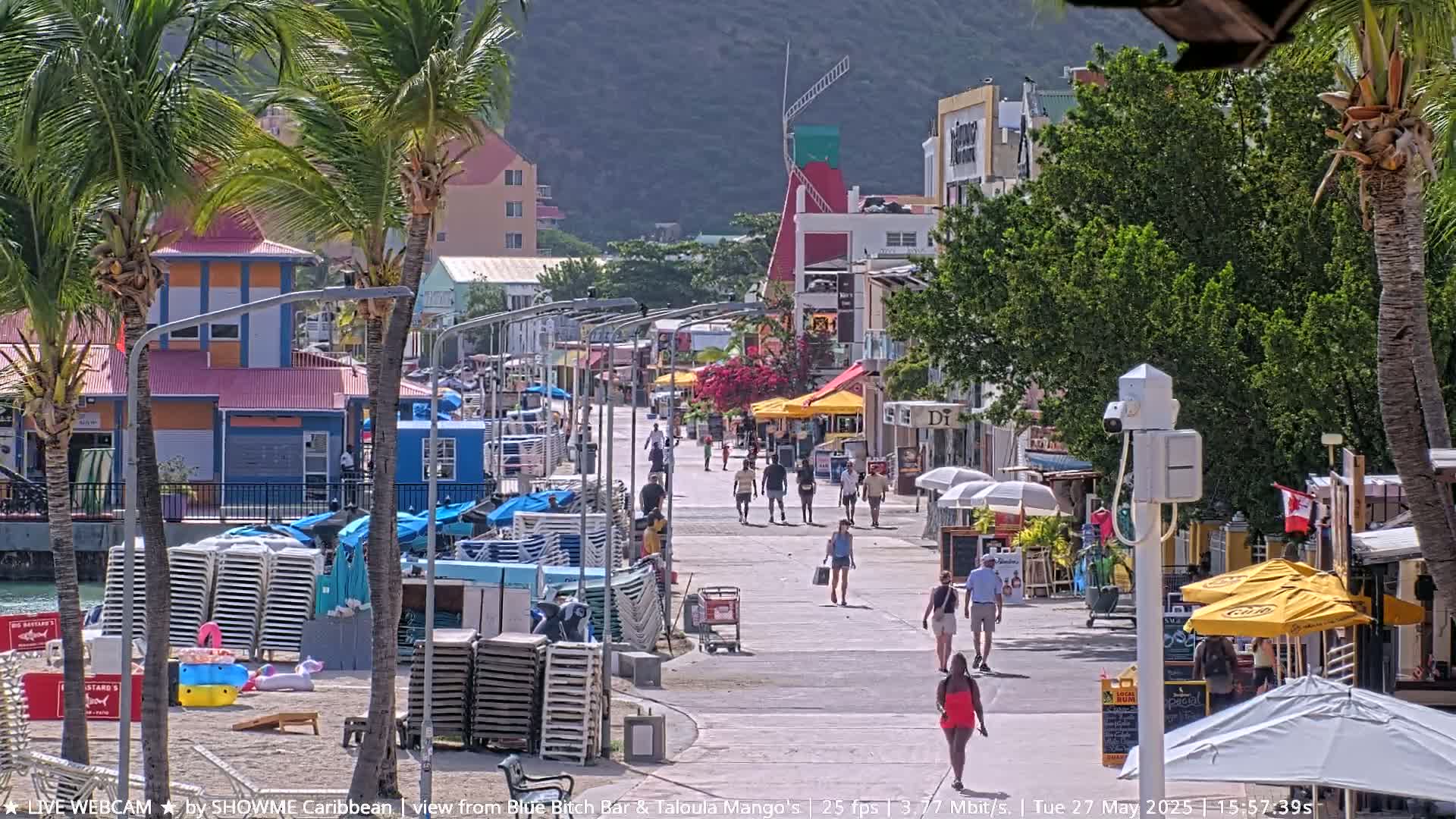 A sunny boardwalk scene shows people strolling past shops and restaurants under palm trees, with beach chairs stacked nearby.