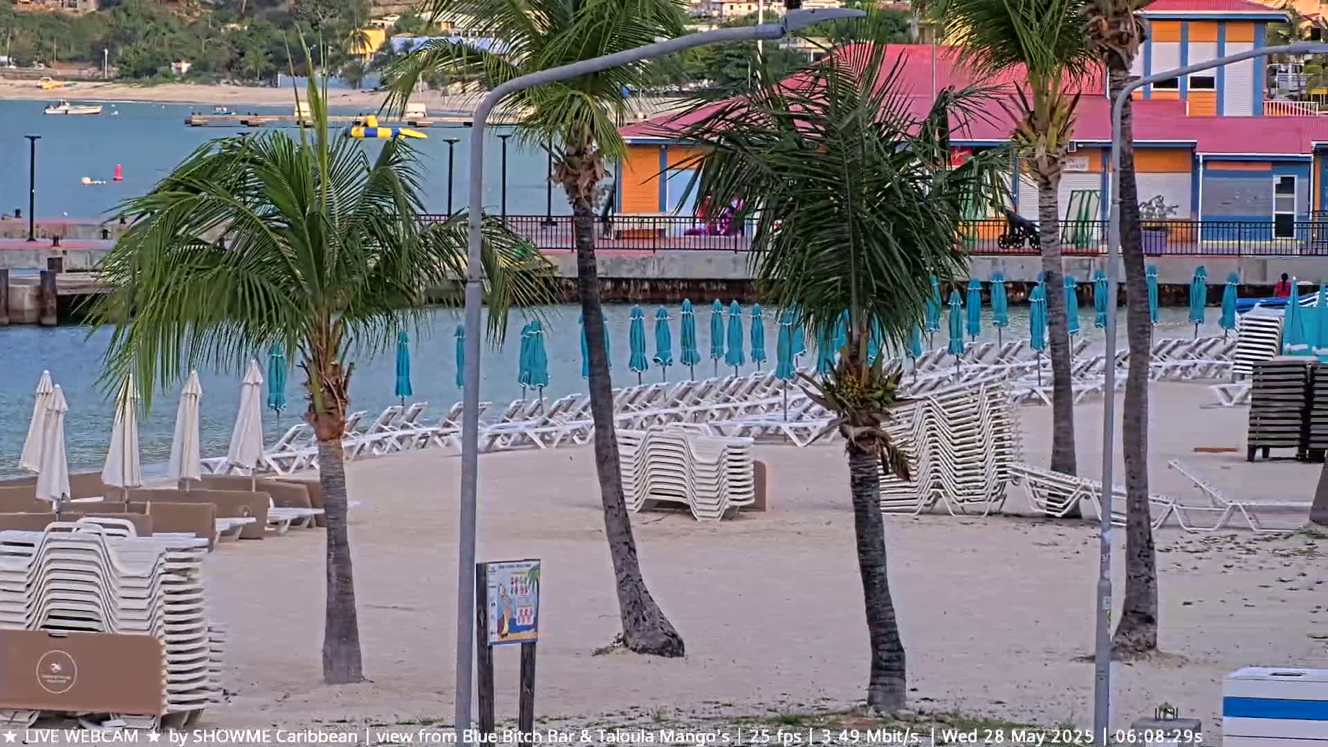 A sandy beach with palm trees, numerous lounge chairs stacked and unstacked, and teal umbrellas is visible, along with calm water and buildings in the background.