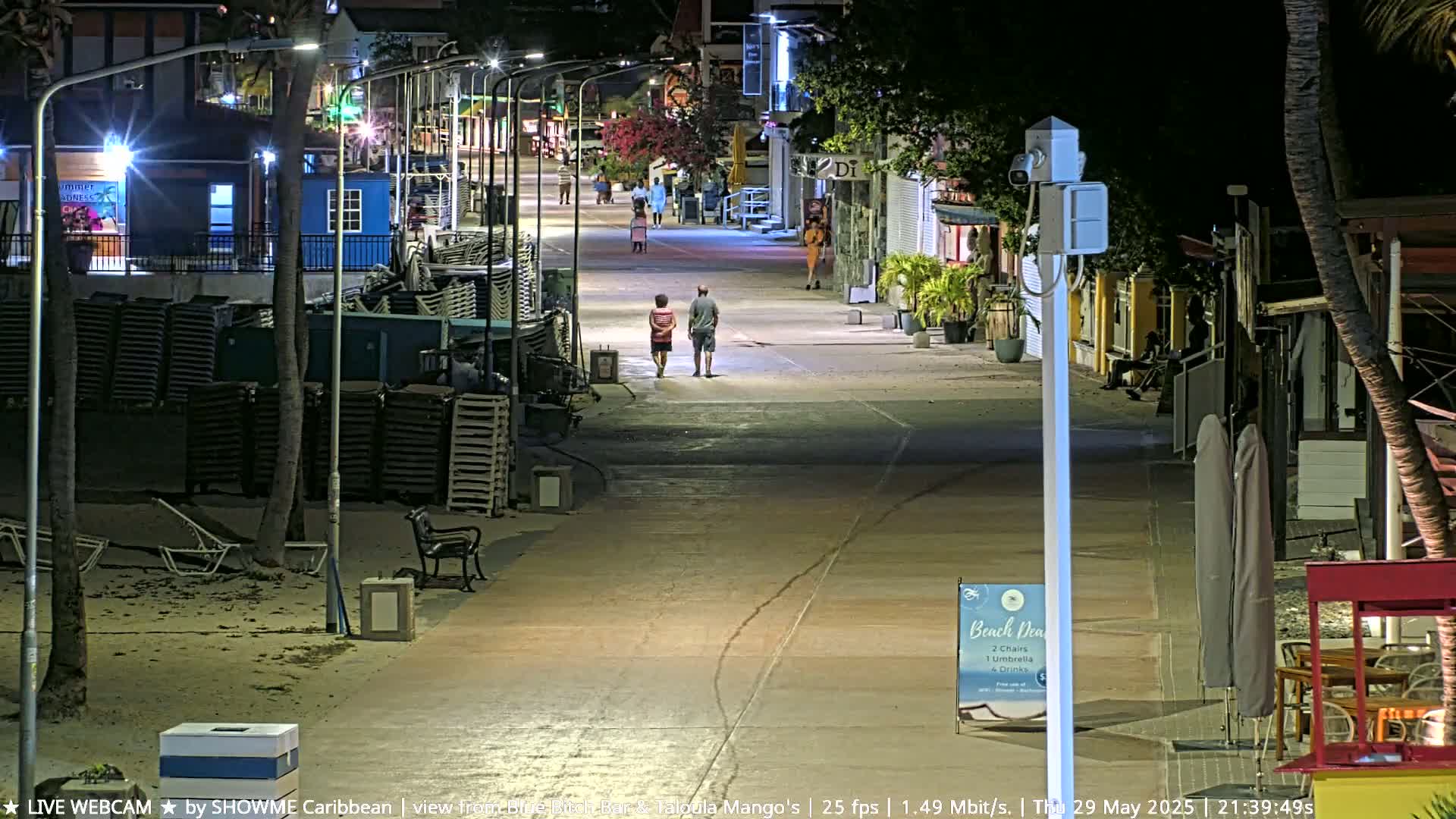 A mostly empty, brightly lit pedestrian street at night, lined with shops and palm trees, with two people walking and stacks of beach chairs along the side.