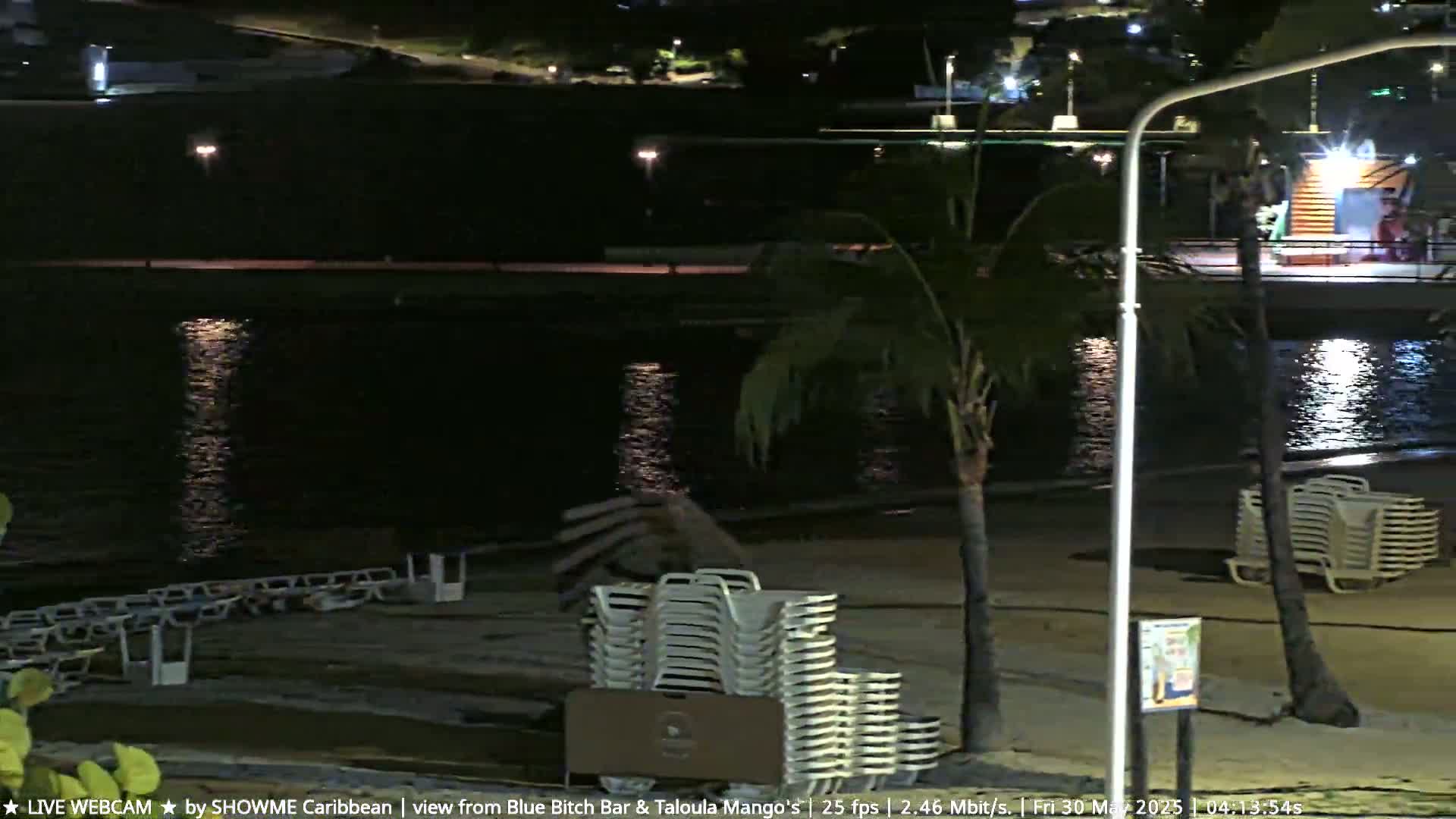 A nighttime beach scene shows calm water reflecting lights, stacks of beach chairs on the sand, and palm trees under a clear sky.