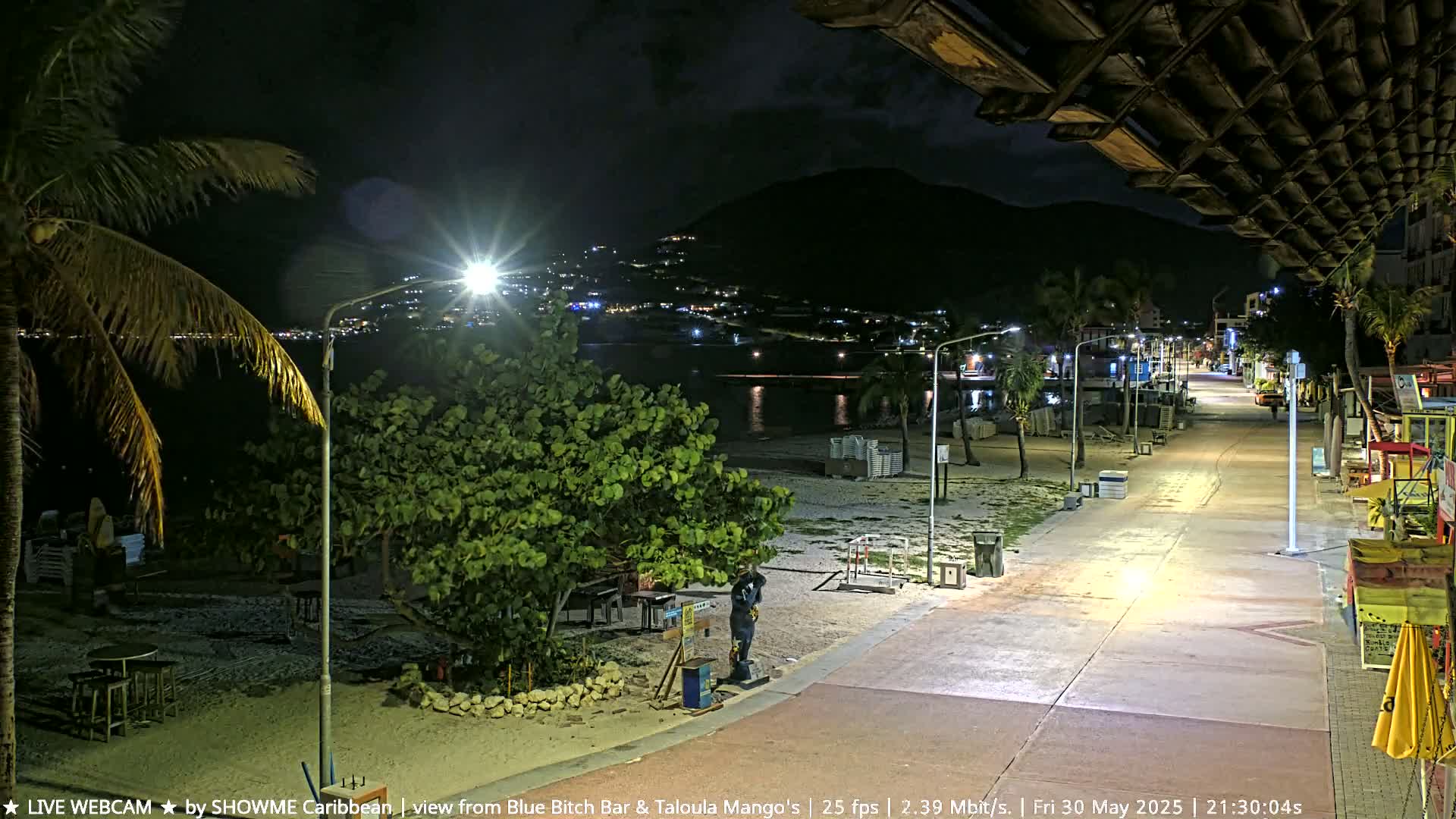 A nighttime beach boardwalk scene shows a mostly empty sandy beach, palm trees, streetlights, and a few buildings in the distance under a clear sky.