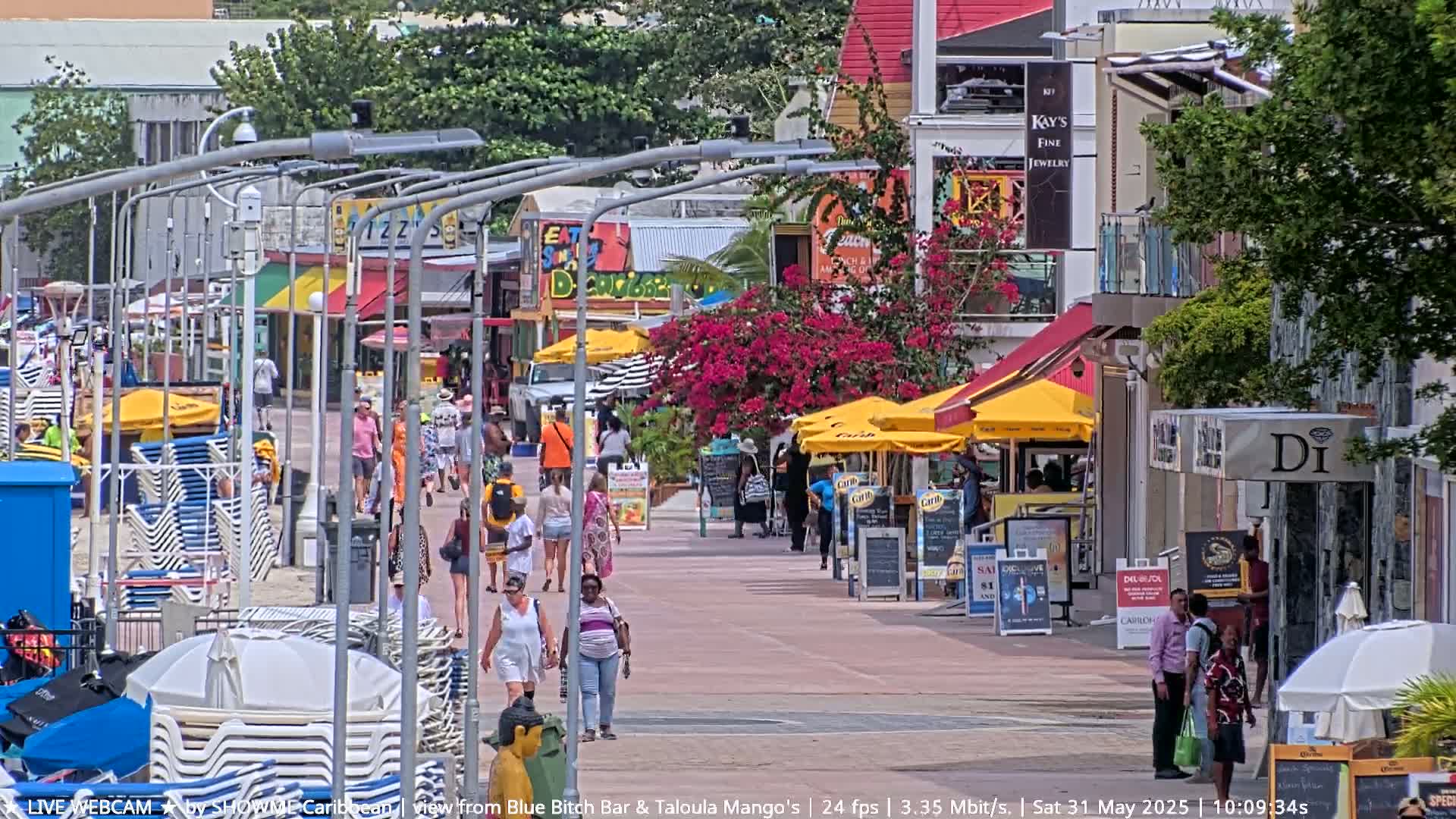 A sunny pedestrian walkway lined with shops and restaurants, populated with people and beach chairs under yellow umbrellas.