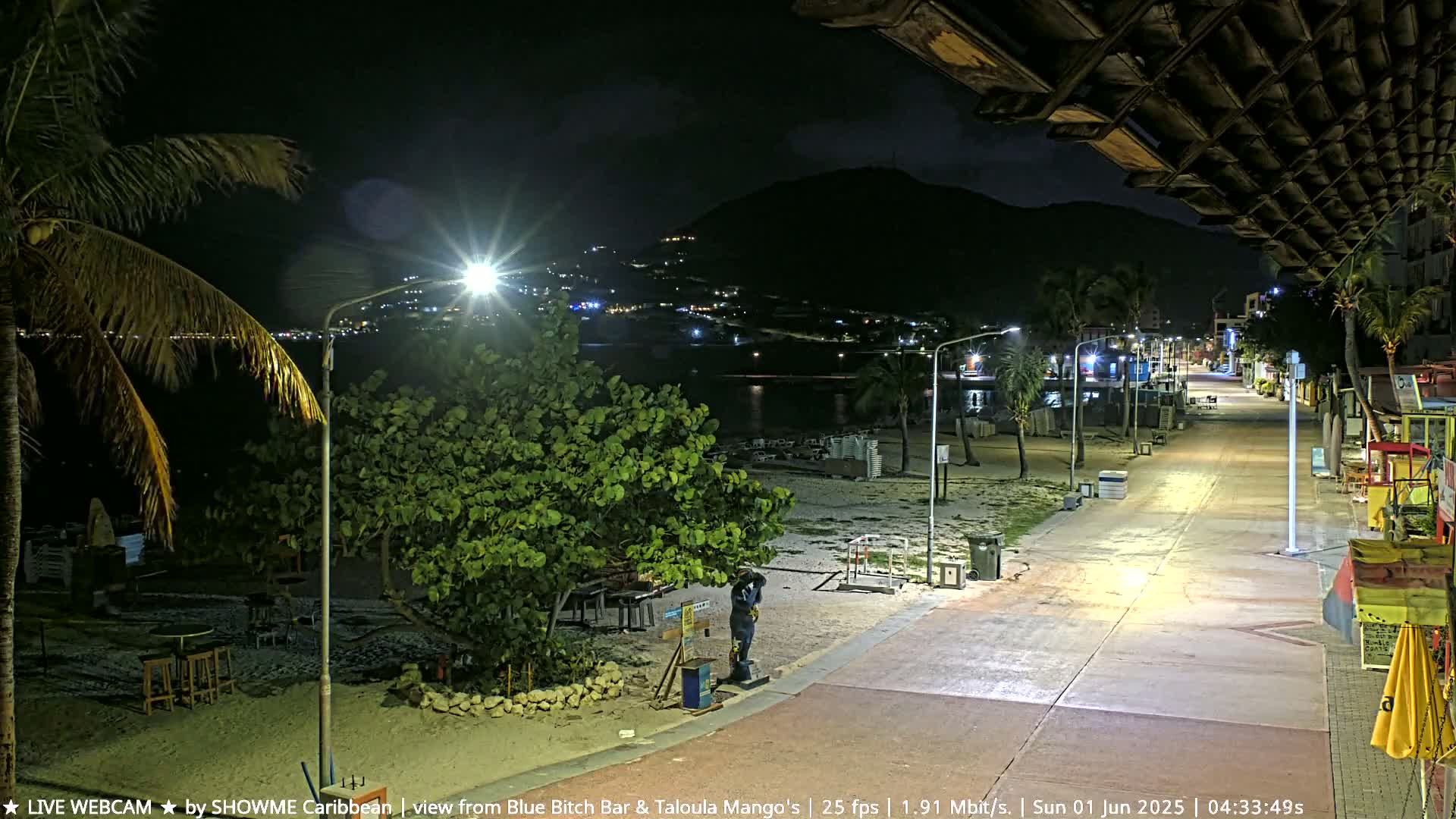 A nighttime view of a mostly empty beachfront boardwalk illuminated by streetlights, with a dark mountain in the background.