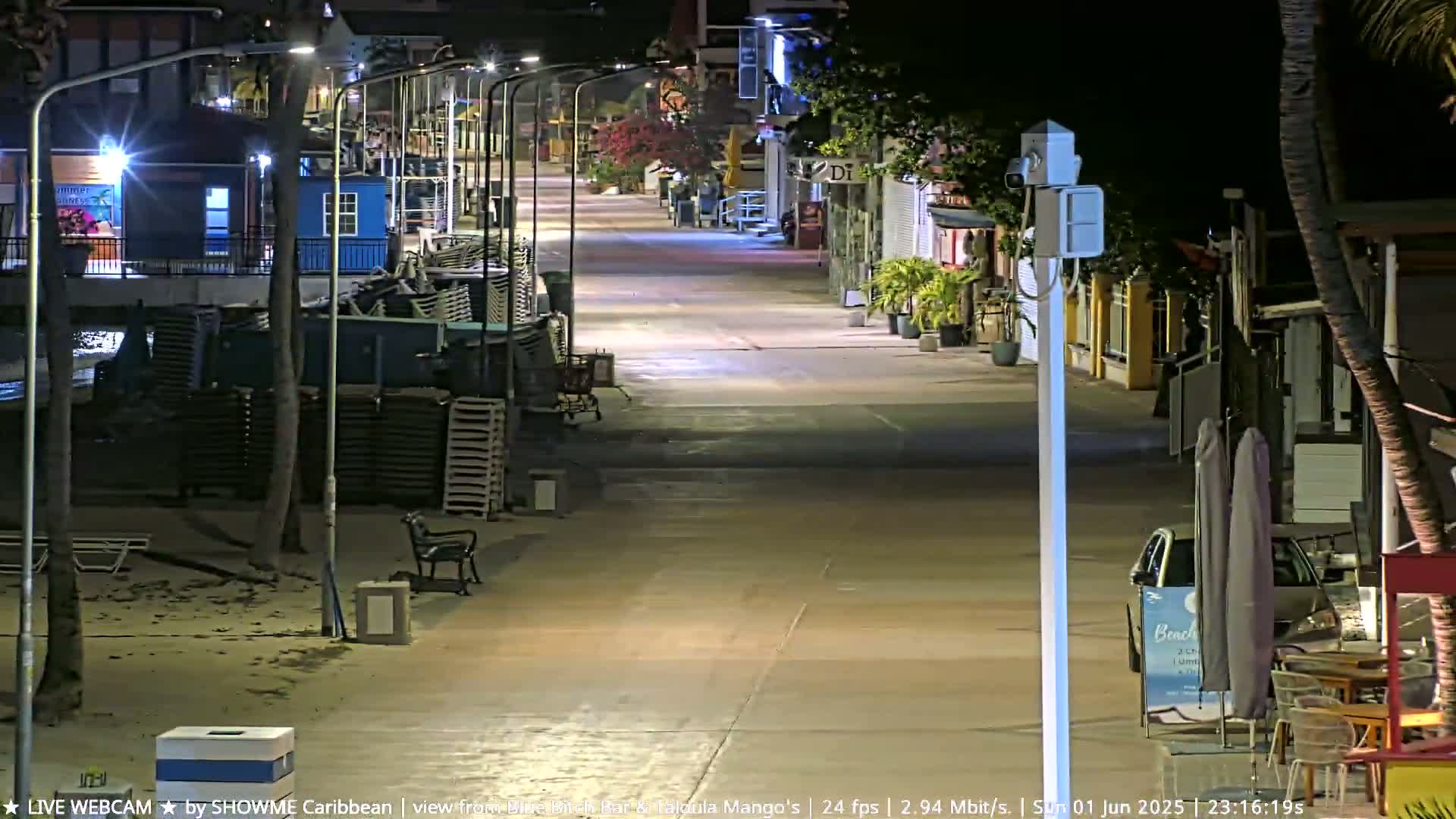A mostly empty, brightly lit beachfront street at night, with stacked beach chairs along the side and a few palm trees.