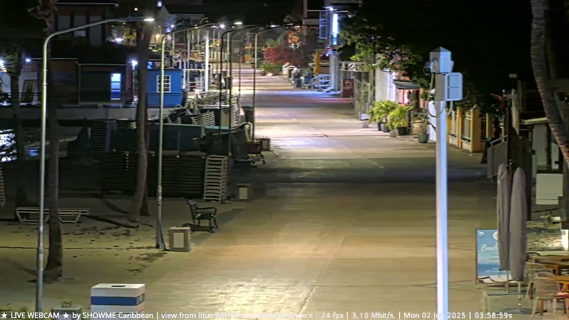 A mostly empty, nighttime street lined with shops and palm trees, next to a sandy beach area.