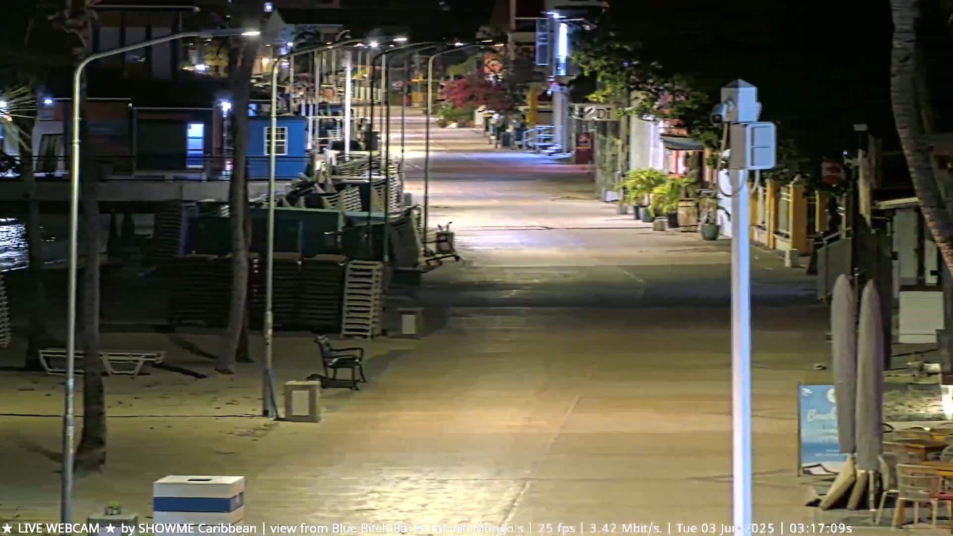 A deserted nighttime boardwalk stretches toward brightly lit buildings, with stacks of beach chairs and a few empty tables visible along the sandy shore.