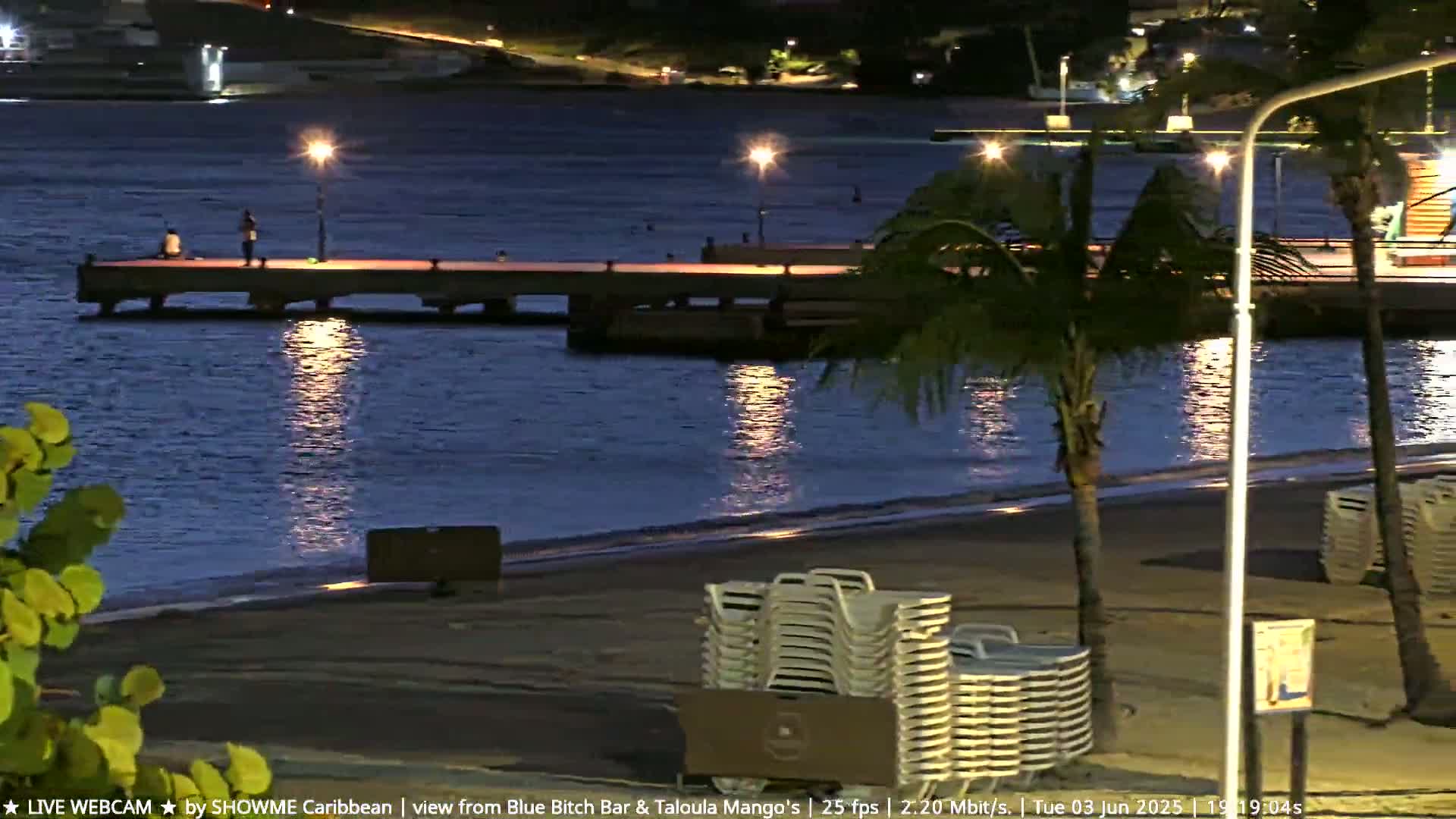 A nighttime beach scene shows a pier with a few people on it, palm trees, and stacked beach chairs on the sand, illuminated by lights along the water.