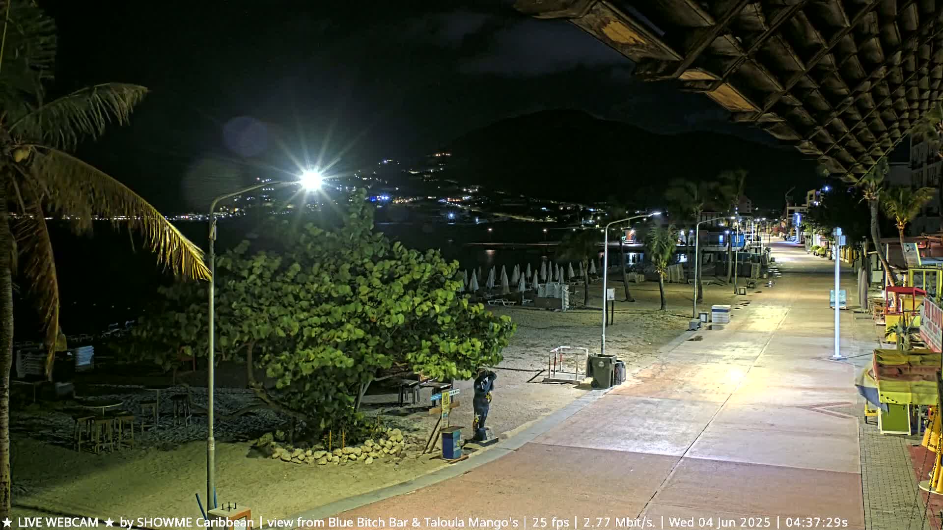 A nighttime view of a beach boardwalk with lights illuminating a mostly empty beach and a few buildings in the distance, under a partly cloudy sky.