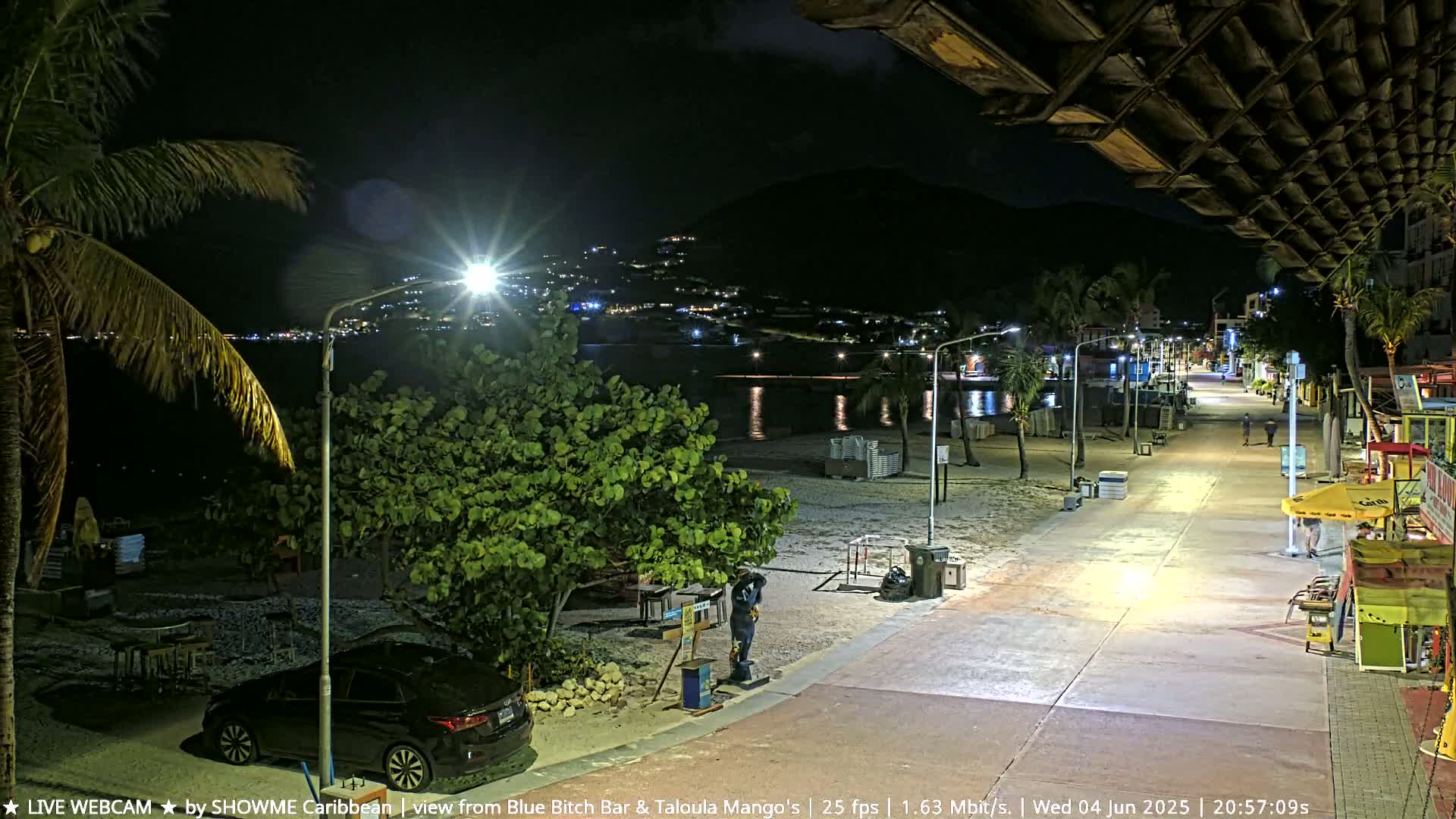 A nighttime beach scene shows a boardwalk lined with palm trees and small shops leading to a sandy beach and calm ocean under a mostly clear sky.