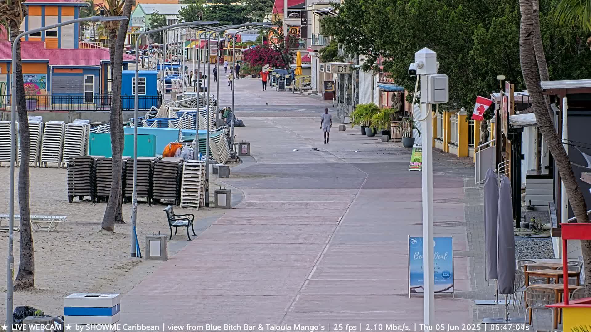 A mostly empty boardwalk runs alongside a beach with stacked beach chairs and palm trees, under a partly cloudy sky.