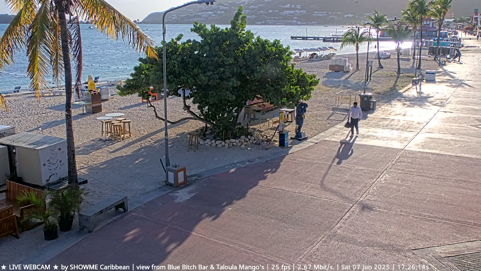 A sandy beach with a few people and outdoor furniture is next to a paved walkway on a sunny day.