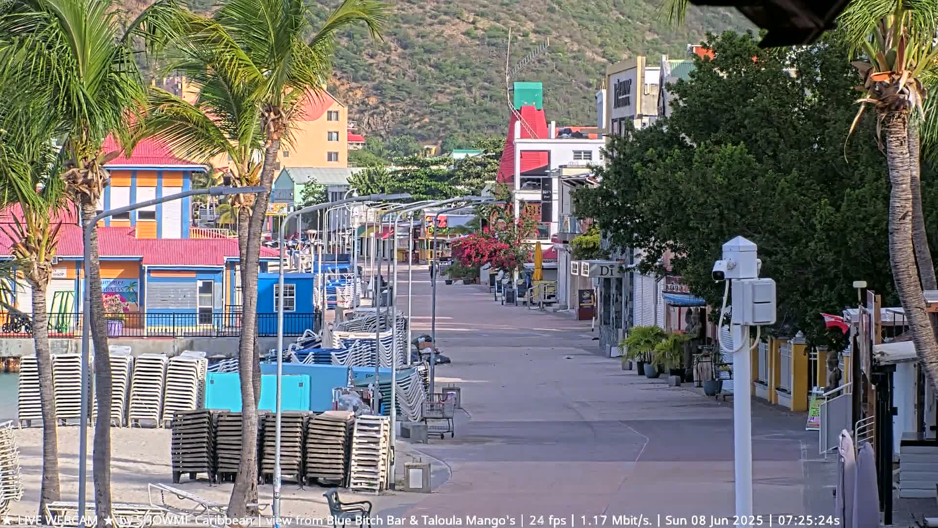 A sunny paved walkway lined with colorful buildings and palm trees leads to a beach with stacked lounge chairs.