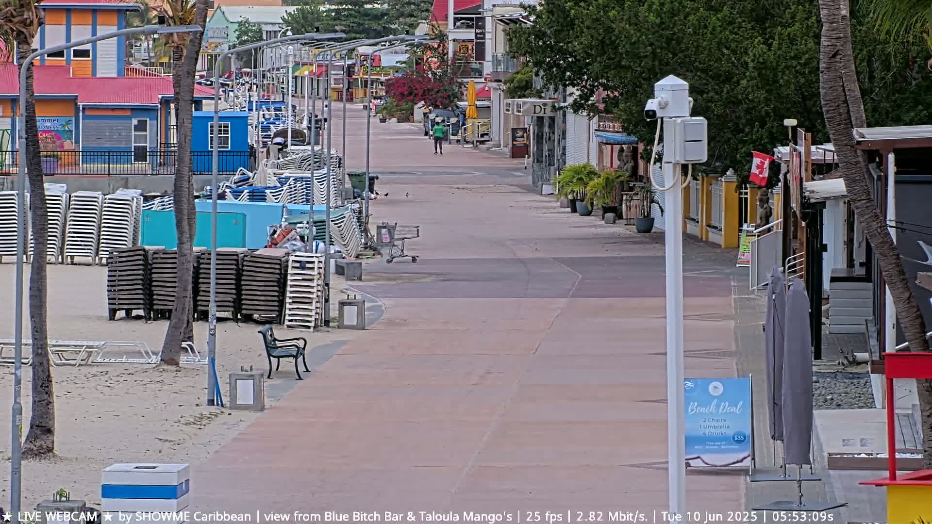 A mostly empty boardwalk on a sunny day, lined with shops and stacked beach chairs, with a few people walking in the distance.