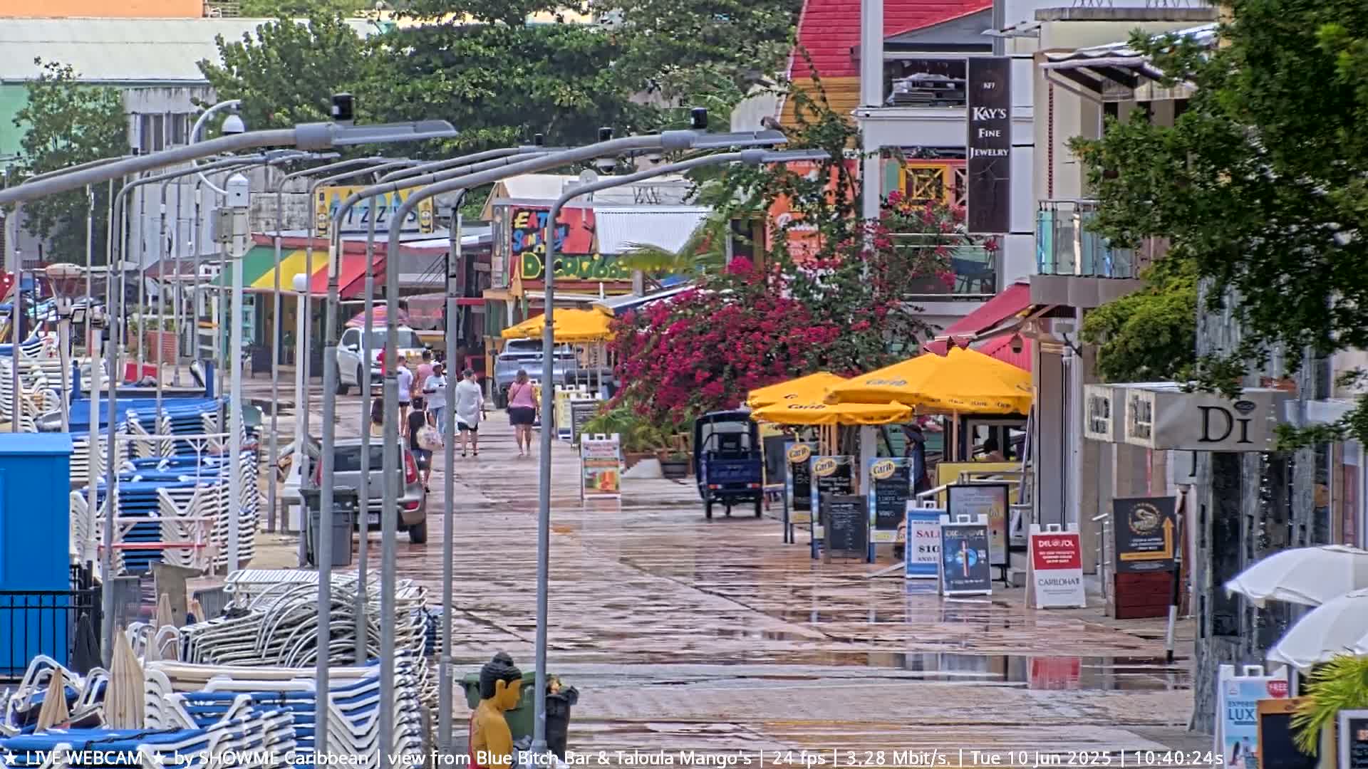A wet pedestrian walkway lined with shops and restaurants, with stacked beach chairs to the side and people walking under a gray sky.