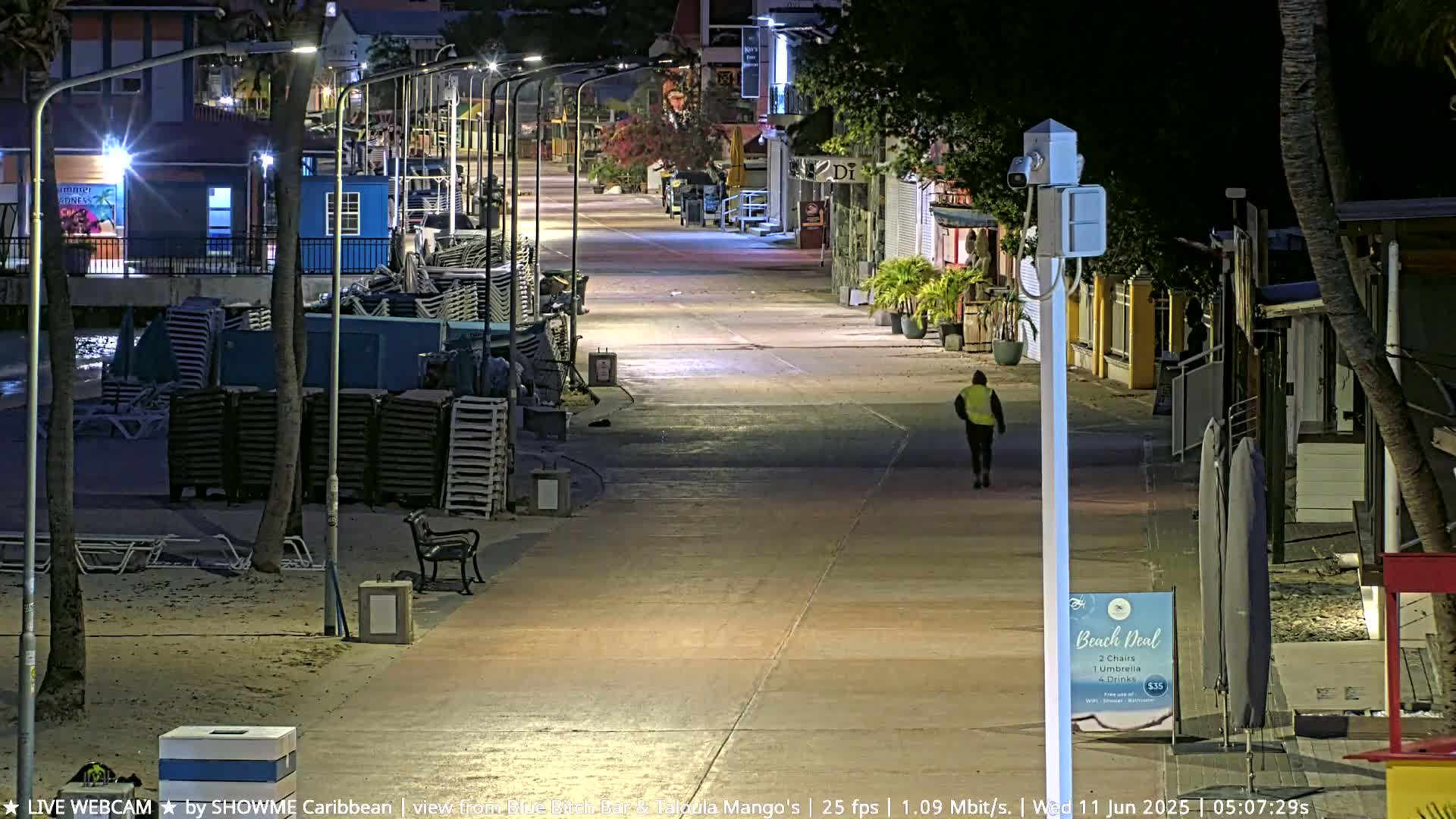 A mostly empty, nighttime boardwalk lined with shops and stacked beach chairs, with a single person walking down the center under bright lights.